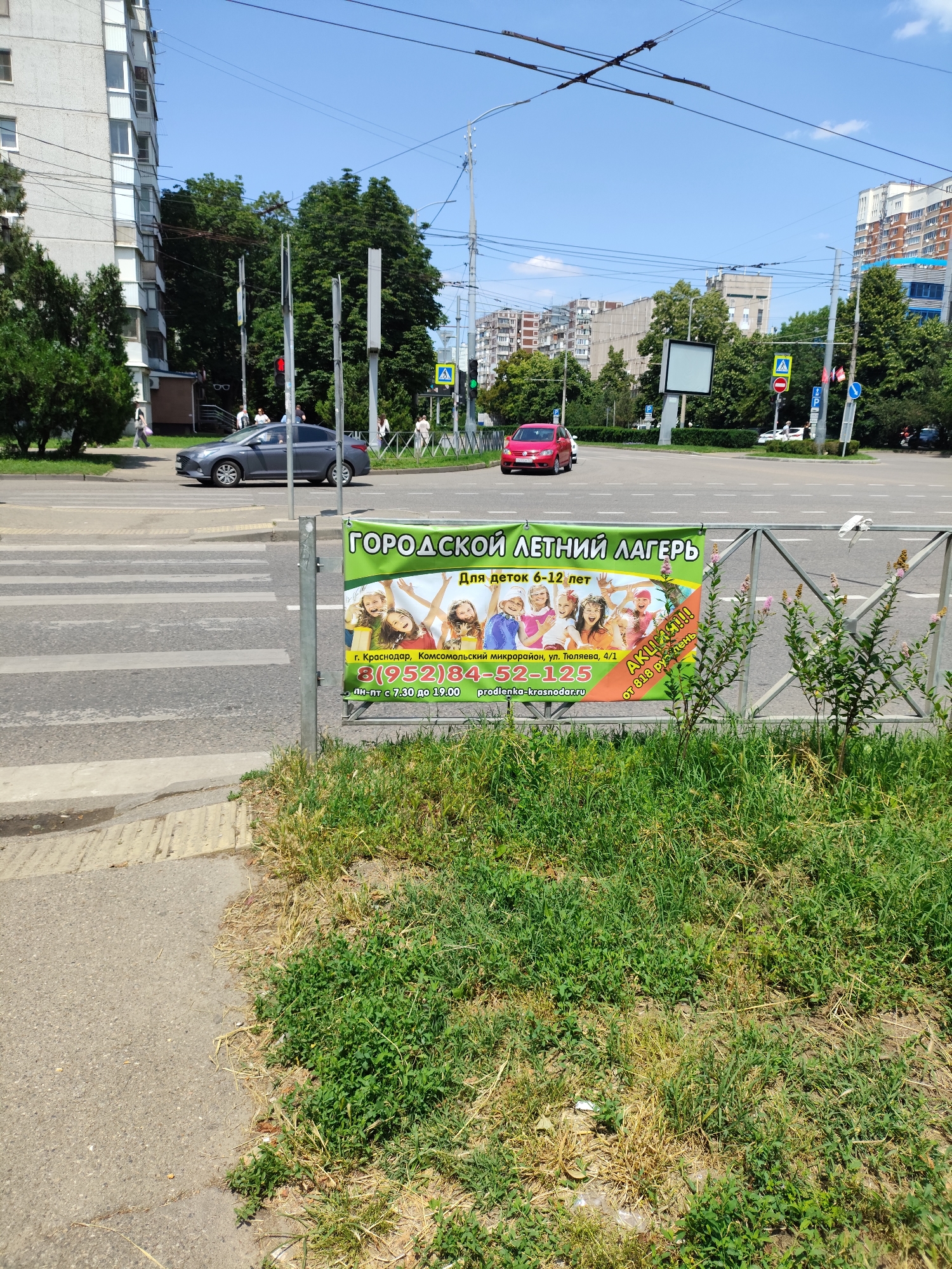 The image captures a sunny day scene in Krasnodar, Russia, focusing on a street intersection with a pedestrian crossing in the foreground. A vibrant green banner advertising a "CITY SUMMER CAMP FOR CHILDREN AGED 6-12 YEARS" is prominently displayed on a metal fence. The banner features smiling children and contact information, including a phone number 8(952)84-52-125 and a website prodlenka-krasnodar.ru. To the left of the banner, a gray sedan is in motion crossing the street. In the background, multi-story buildings, lush green trees, and overhead power lines are visible, along with other vehicles and pedestrian activity. The environment appears to be a typical urban setting during daylight hours.