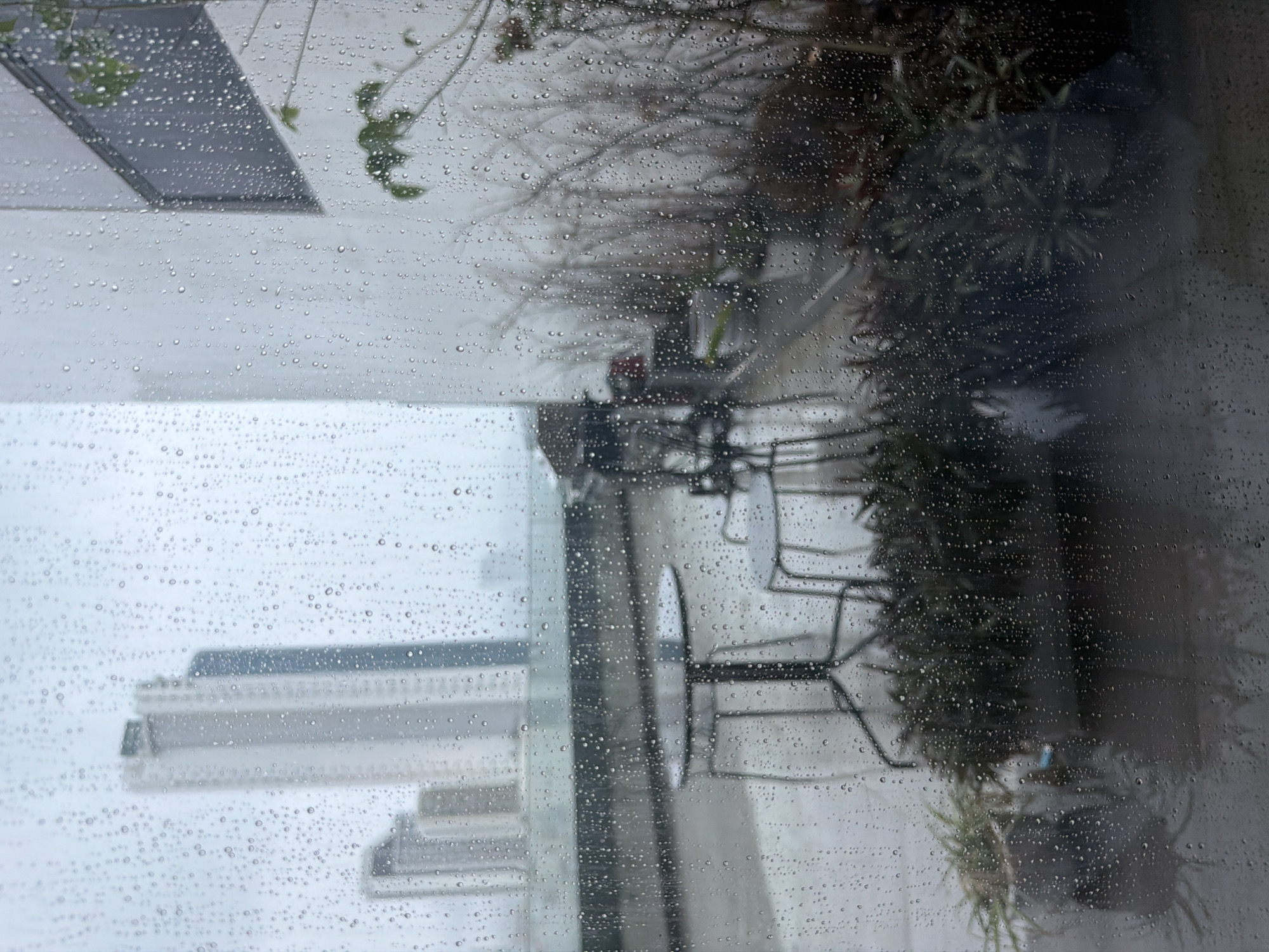 The image shows a view through a rain-streaked window, likely taken from an elevated position, potentially in Bac Nha Trang Ward, Vietnam. Outside the window, a building with multiple horizontal sections is visible, shrouded in mist. On what appears to be a balcony or patio, there are chairs and what looks like potted plants. The rain droplets on the window obscure and distort the view, creating a sense of enclosure.