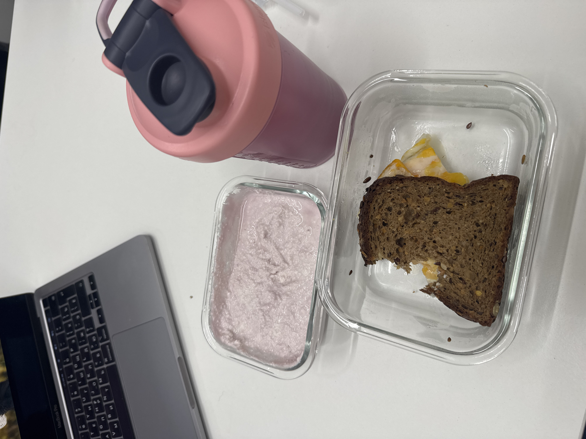 A high-angle shot shows a MacBook Pro, a pink water bottle, and two glass food containers on a white surface. The laptop is partially visible in the lower-left corner, displaying a keyboard and trackpad. The pink water bottle sits to the upper-left of the frame. To the right of the bottle, two clear glass containers hold food items. One container holds a pink, smooth substance, while the other contains a piece of dark bread with an egg. The overall arrangement suggests a lunch or workspace setting. The scene appears to be indoors, possibly an office or home environment, based on the surface and objects present. This location is unlikely to be in Dubai, United Arab Emirates, as the objects are commonplace and provide no indication of the city.