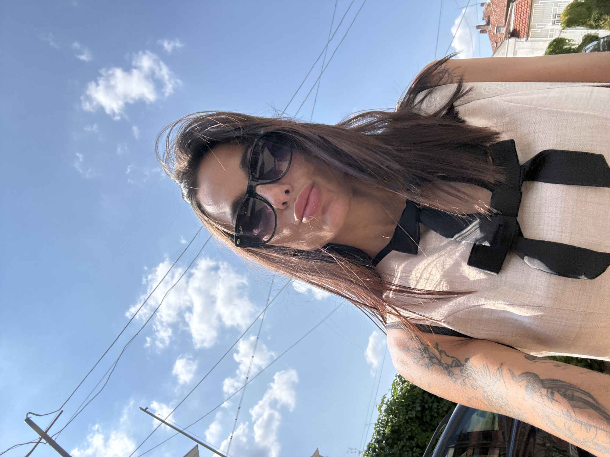 This is a close-up, selfie-style photograph of a woman taken outdoors on a bright, sunny day. The woman, who appears to have long, dark brown hair, is wearing sunglasses and a light-colored dress with a black bow detail at the collar. Her arm, visible in the lower right of the frame, has multiple tattoos.
The background consists of a clear blue sky with scattered white clouds. Several power lines crisscross the sky, and in the upper right corner, the rooftops of buildings, possibly in a European style with red tiles, can be seen. Green foliage is visible in the lower right portion of the image, suggesting a vegetated area.
The lighting indicates it is daytime, likely late morning or afternoon, given the strong sunlight casting shadows and highlighting the woman's features. The overall mood appears casual and relaxed.
No visible text is present in the image. The setting suggests an urban or suburban environment, possibly a street or garden area. The photograph was taken in Pleven, Bulgaria.
