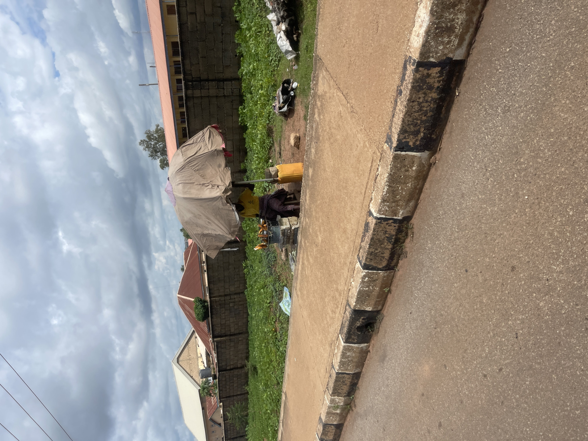 A person is selling corn on the cob under a large umbrella on a dirt and grassy area next to a sidewalk in Barakin Lerawi, Nigeria. The corn is being roasted over a fire in a metal bucket. A yellow container sits to the right of the person. In the background, there is a brick wall and buildings. The sky is cloudy.