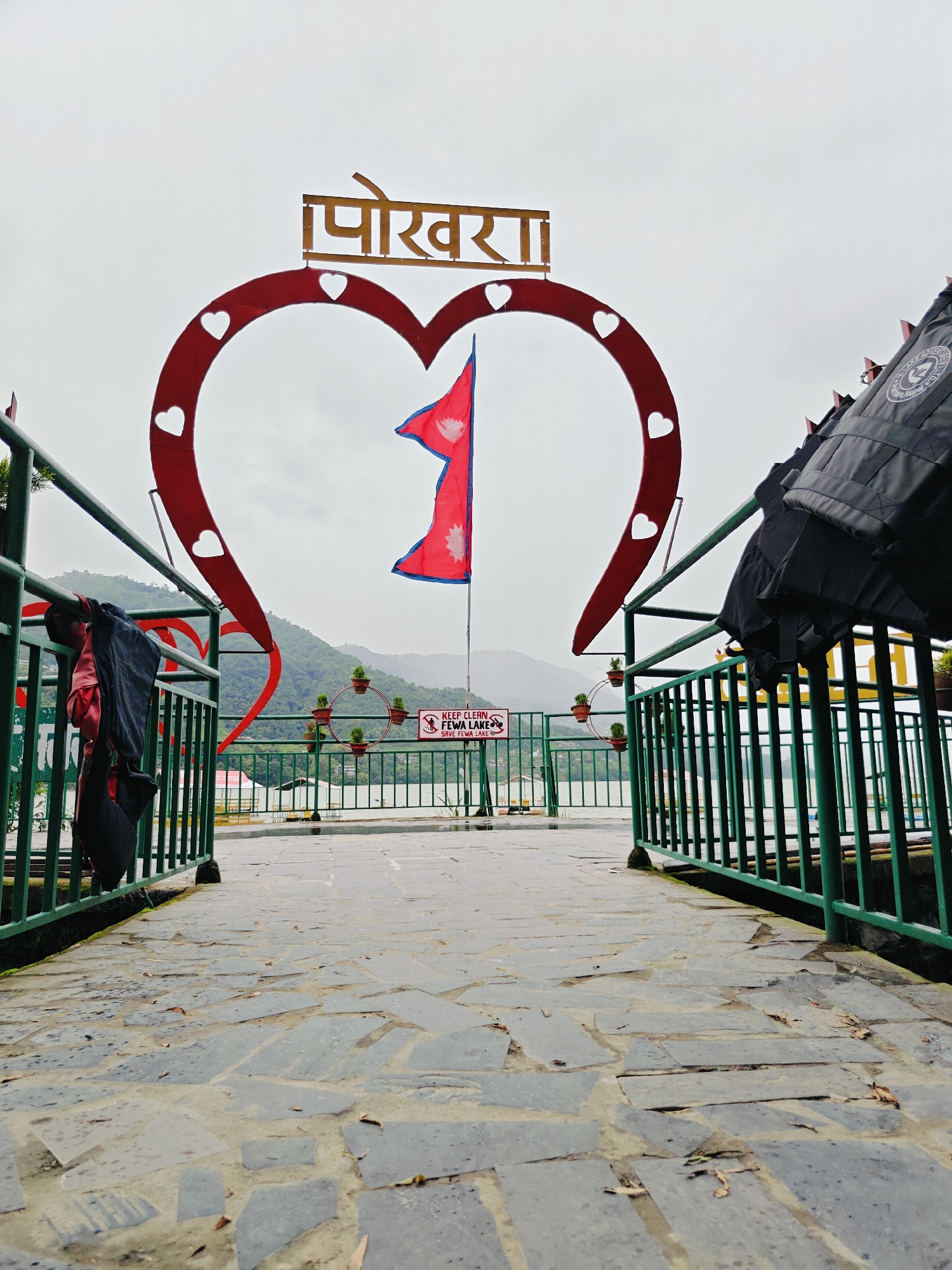 The image depicts the entrance to Phewa Lake in Pokhara, Nepal, marked by a large heart-shaped archway with the word "पोखरा" (Pokhara) inscribed in Devanagari script at the apex. A Nepalese flag is positioned in the center of the arch, fluttering gently. The arch is red and adorned with smaller white heart cutouts. To the left and right of the archway, green metal railings line a paved walkway. Several life jackets are hung on the left railing, and a sign on the right railing reads "KEEP CLEAN THE FEWA LAKE SAVE FEWA LAKE". In the background, the calm waters of Phewa Lake are visible, with lush green hills and mountains under an overcast sky. The pathway in the foreground is constructed from irregular gray stone slabs, with scattered fallen leaves indicating a natural outdoor setting. The overall scene suggests a popular tourist spot, likely a viewpoint or entrance area for activities on the lake.