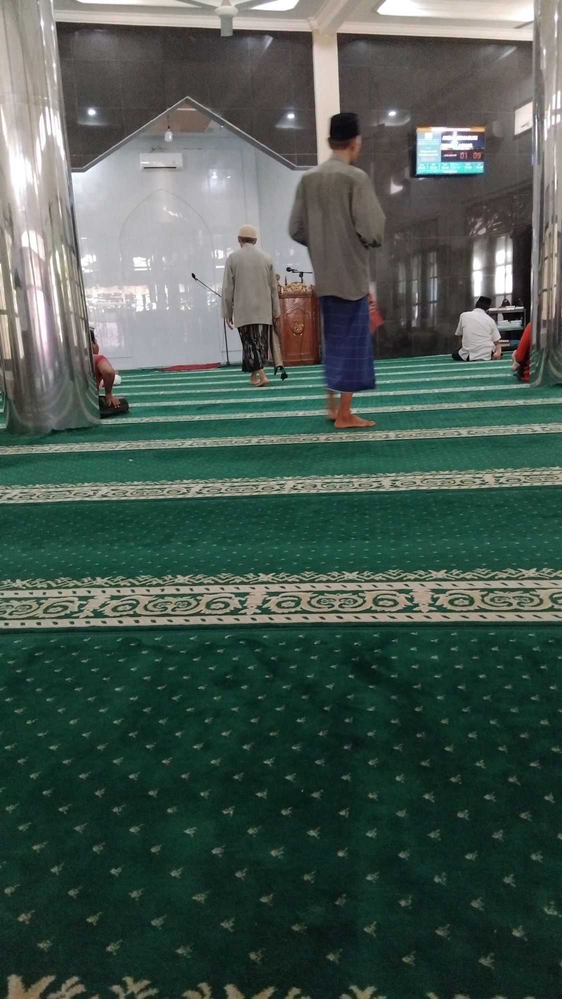 A group of people are inside a mosque, with green carpeting and white patterned borders laid out in rows. In the foreground, the carpet is a deep green with a subtle, repeating small diamond pattern. Further back, the carpet has wider white borders with intricate scrollwork designs.
Two men are standing in the middle of the frame. The man on the right, wearing a dark blue sarong and a grey long-sleeved shirt, faces away from the camera. He has a black skullcap on his head. The man to his left, also facing away, wears a light-colored sarong with a dark pattern and a light grey long-sleeved shirt, and a white skullcap. Another man in a white shirt and dark pants is seated on the floor to the right, facing the wall. A red-shirted individual is partially visible on the far left, kneeling.
In the background, a large speaker stands on a wooden podium. Above the podium, a digital display shows the time "01:09". The mosque features white walls and a vaulted ceiling, with a light fixture visible in the upper center. Large, polished metal pillars flank the sides of the image. The scene appears to be the interior of a mosque in Batang, Indonesia.