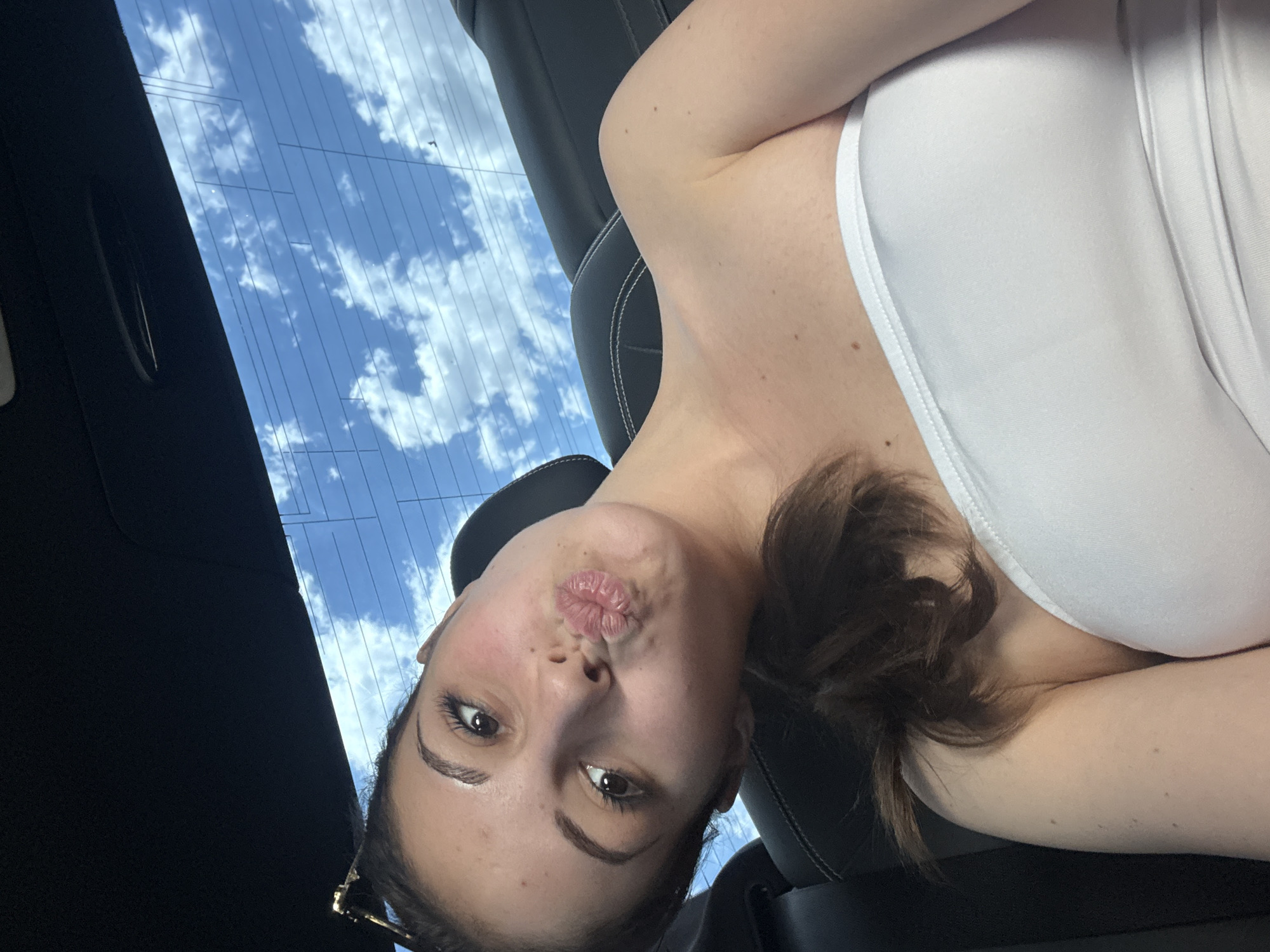 A young woman with brown hair is taking a selfie in the back seat of a car. She is wearing a white strapless top and has her lips pursed. The background shows the sky through the car's rear window. The image does not provide any information about the location being Burgas, Bulgaria.
