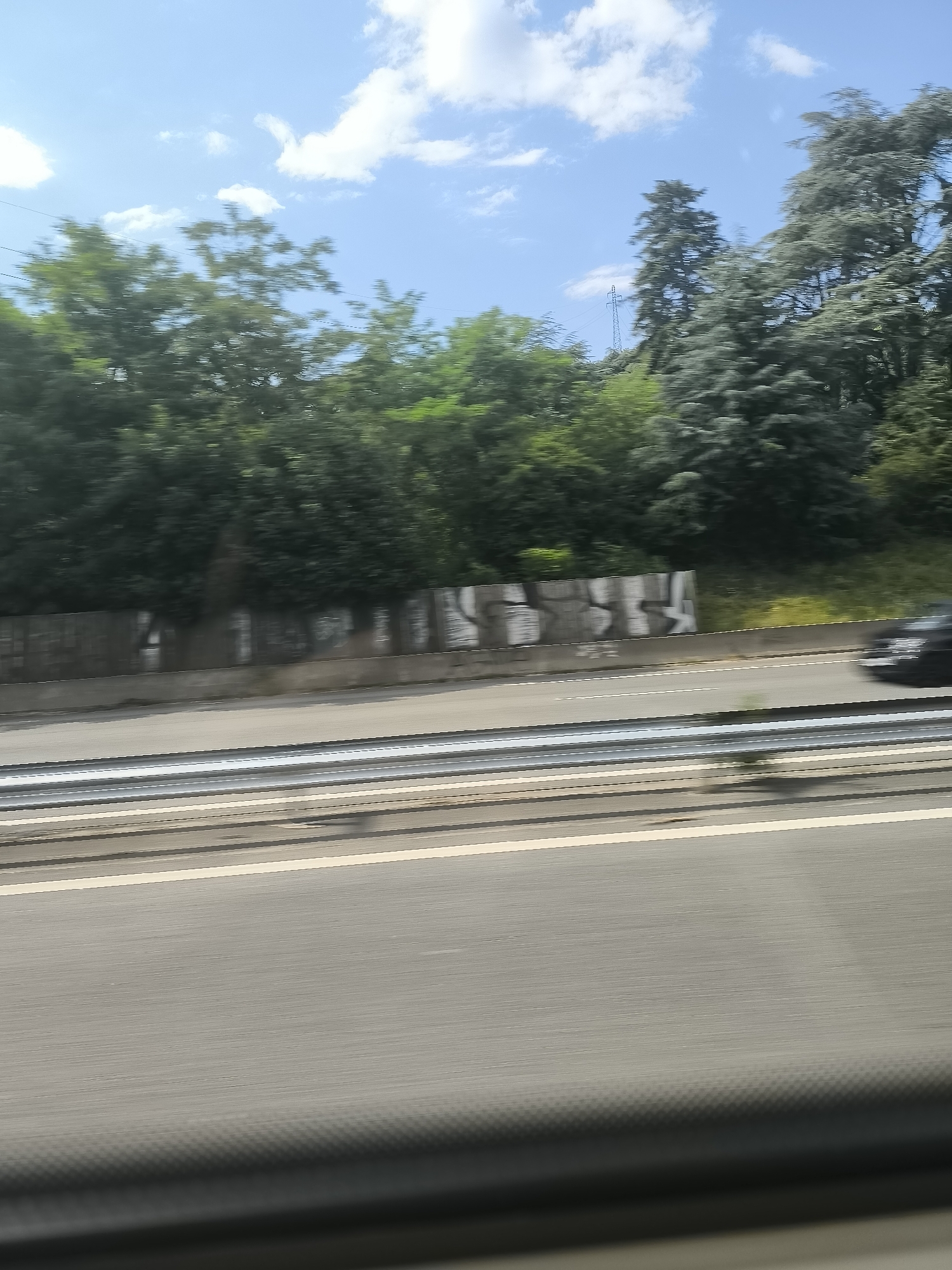 The image, likely taken from a moving vehicle, shows a highway landscape on a sunny day in Saint-Jean-Bonnefonds, France. The road occupies the lower portion of the frame, showing several lanes with painted white lines and a barrier. Beyond the barrier is a concrete retaining wall covered in graffiti. Above the wall is a line of dense trees and vegetation. In the distance, partially obscured by trees, is a power line pylon. A dark-colored vehicle is partially visible on the right side of the highway. The sky is blue with scattered white clouds.
