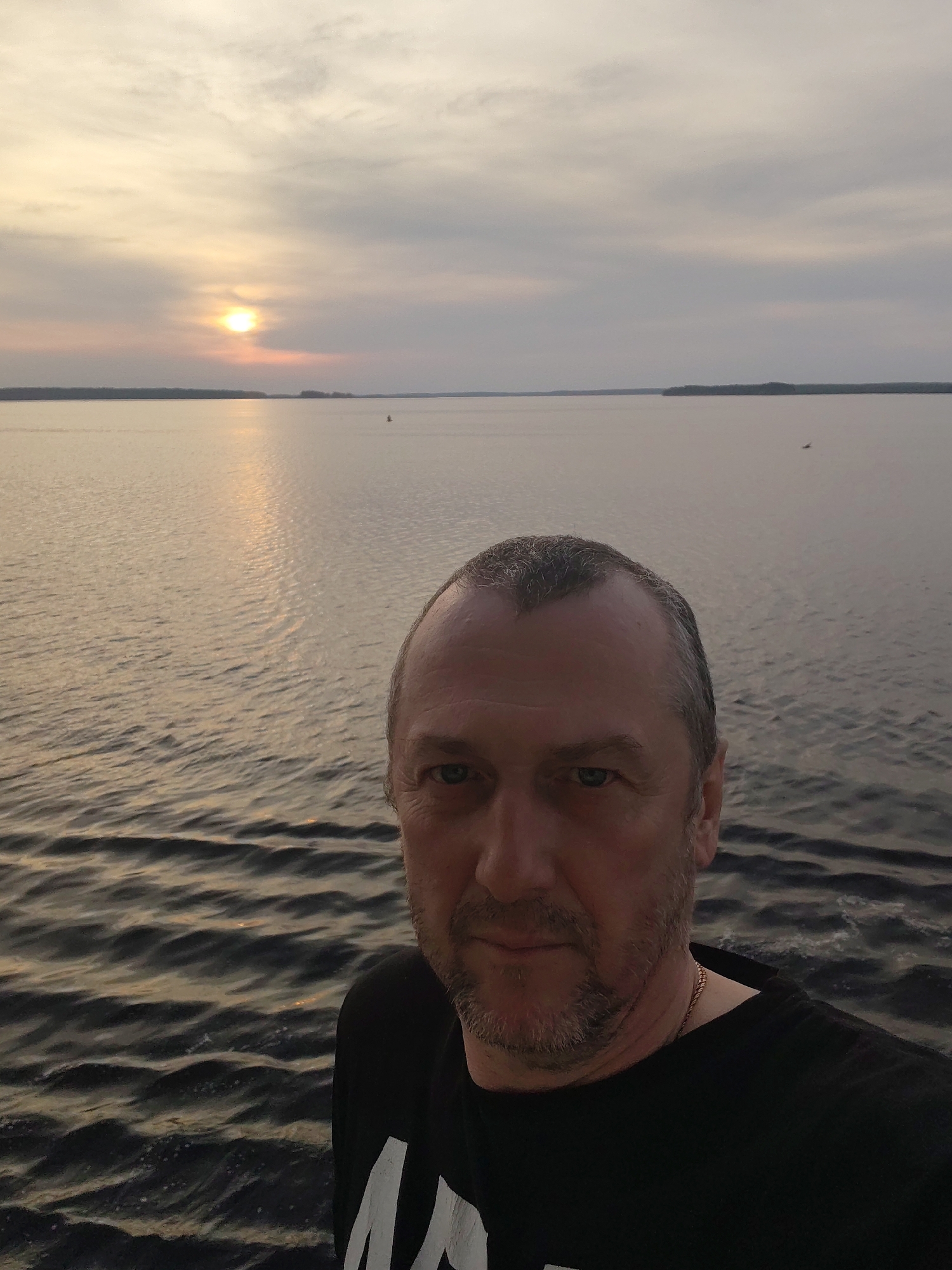 A selfie photograph taken in what appears to be Суда, Russia at sunset. The foreground contains a man wearing a black t-shirt and gold chain, centered and facing the camera. The background shows a calm body of water, presumably the Suda River or a nearby lake, reflecting the setting sun. The sky is overcast with clouds. There is land visible on the horizon.