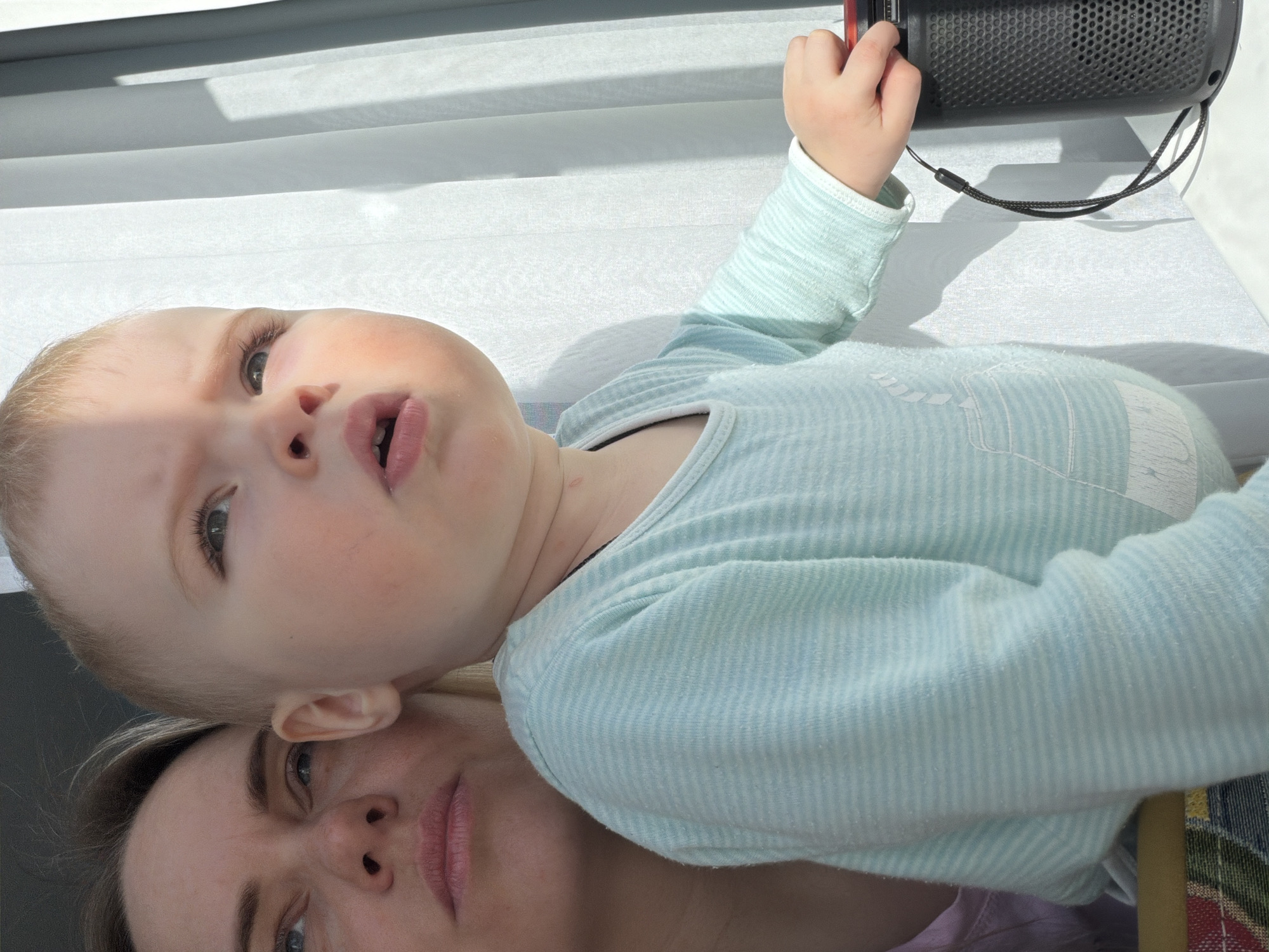 The image portrays an indoor scene in Votkinsk, Russia, illuminated by natural light. In the foreground, a young child, appearing to be a toddler, is depicted from the upper chest upwards. The child possesses light-colored hair and fair skin, with their gaze directed upwards and to the right of the frame, and their mouth slightly agape. They are attired in a light blue, long-sleeved top featuring thin white horizontal stripes and a subtle white graphic on the lower front. A dark cord or necklace is visible around the child's neck. The child's right hand rests upon a dark, cylindrical, perforated object consistent with a portable speaker, which has a red-colored top accent and an attached black lanyard, positioned on a white surface.
Behind and slightly to the left of the child, a woman's face is partially visible, from the nose upwards. She has light-colored hair and skin, and her eyes are also directed upwards and to the right, aligning with the child's gaze. The background to the right features white, vertical blinds or sheer curtains, displaying distinct shadow patterns caused by direct sunlight streaming into the space. A glimpse of multi-colored patterned fabric is visible in the lower-left corner of the frame.