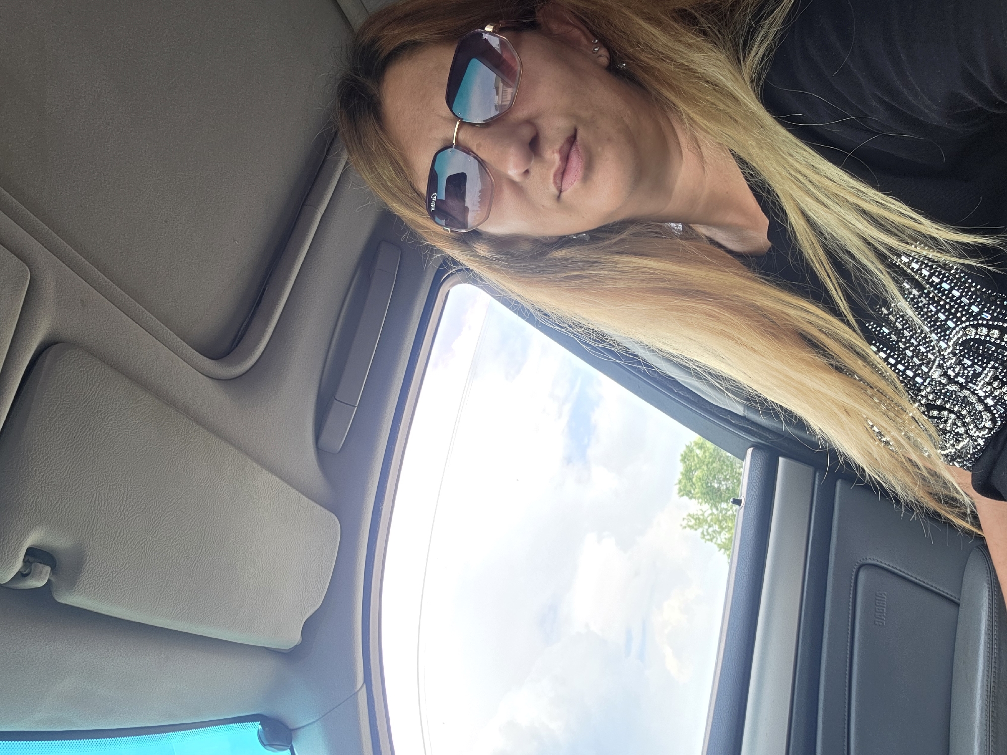 A medium-shot photograph taken inside a vehicle during daylight. The subject is a woman with blonde hair wearing sunglasses and a dark-colored, possibly black, top with embellishments. The woman is looking at the camera. The car interior includes a gray roof, sun visor, and door panel. The window shows a view of the sky and a tree, suggesting an outdoor environment. The photograph was taken in Varna, Bulgaria.