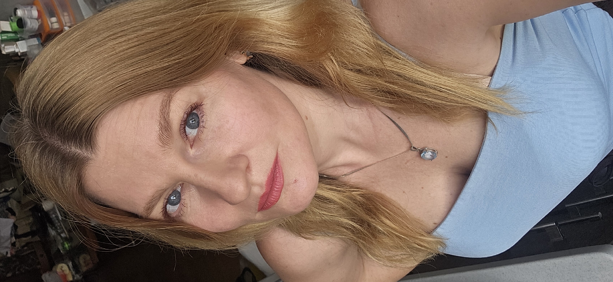 The image shows a close-up, high-angle portrait of a fair-skinned woman with blonde hair and blue eyes. She is wearing a light blue top and a silver necklace with a light blue pendant. Her lips are painted with a coral-colored lipstick. The background is out of focus and appears to be an indoor setting with shelves holding various bottles and containers. The lighting suggests it is daytime. The image does not contain any specific visual cues that would indicate its location within the city of Krasnodar, Russia.