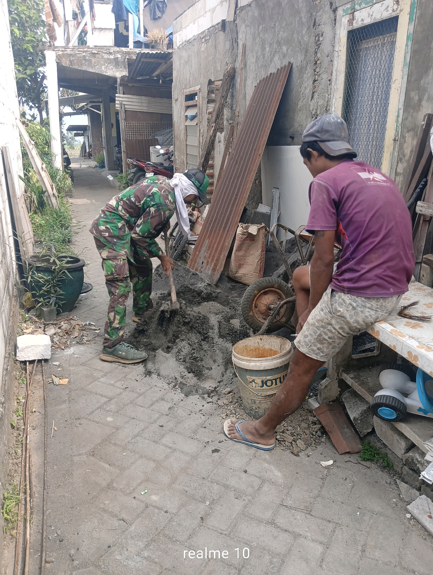 In an outdoor, narrow alleyway setting in Batu, Indonesia, two individuals are engaged in a construction-related activity. One person, wearing camouflage pants and a camouflage top with a white head covering, is actively mixing cement on the ground with a shovel. They are wearing green athletic shoes. The other person, wearing a purple t-shirt and patterned shorts, is seated on a makeshift bench made of concrete blocks and a patterned surface, observing the activity. This person is wearing a grey baseball cap and flip-flops.
The environment is characterized by a mixture of paved brick flooring and loose gravel. Various construction materials are visible, including a bag of cement, a wheelbarrow, a large plastic bucket labeled "JOTUN," and stacks of corrugated metal sheets and other building debris. The buildings flanking the alleyway appear to be constructed from rough, unfinished concrete, with some areas showing exposed brickwork. Laundry is hung on lines between buildings in the background, suggesting a residential area. The lighting suggests it is daytime, possibly late morning or afternoon, with no strong shadows indicating direct overhead sun. The overall impression is a scene of manual labor in a functional, somewhat unkempt urban environment.