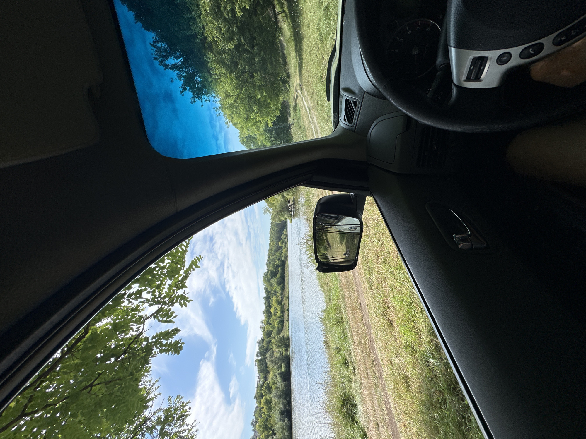 The image is a first-person view from inside a car, looking out the driver's side window and through the windshield. The car is parked or driving slowly on a grassy bank next to a wide body of water, likely a river. Lush green trees and vegetation line the opposite bank and extend up the rolling hills. The sky is blue with scattered white clouds. The interior of the car shows the dashboard, steering wheel, and part of the door panel. The side mirror reflects a portion of the scene. The location is identified as Makiv, Ukraine.