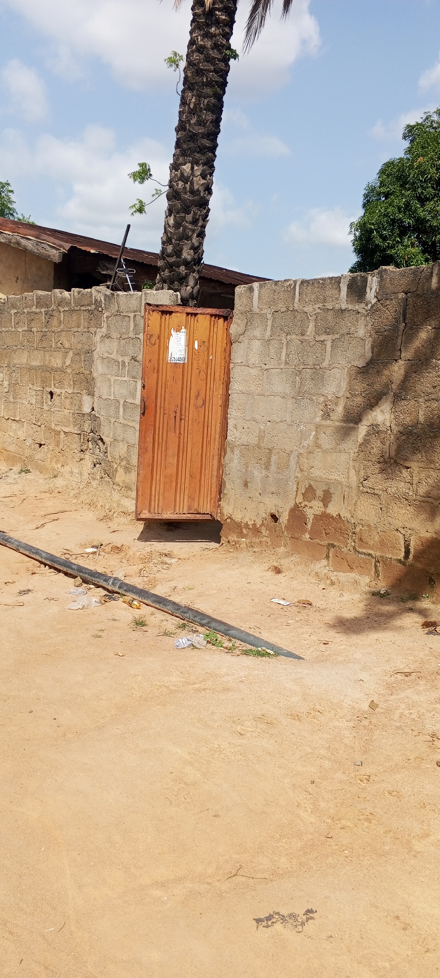 The image depicts an outdoor scene in Jalingo, Nigeria. The focal point is a weathered, orange metal gate set within a textured concrete block wall. The gate has vertical fluting and appears to be slightly ajar. A white sticker with some illegible text and the numbers "253456" is affixed to the right side of the gate.
Behind the wall, a portion of a rusty metal roof is visible, suggesting a building. To the left of the gate, a tall, rough-barked palm tree trunk dominates the upper left quadrant of the image, with a few small green shoots emerging from its sides. A dark, flexible pipe, likely for water, lies on the sandy ground in the foreground, running diagonally from the left towards the bottom right. Scattered debris, including plastic bags and a yellow bottle cap, are visible around the pipe and on the ground.
In the background, a lush green tree with dense foliage stands to the right of the wall. The sky above is a light blue with scattered white clouds, indicating fair weather. The sandy ground suggests a dry or arid environment. The overall impression is one of an ordinary day in a residential or utilitarian area, possibly in the afternoon given the shadows. There are no people visible in the image.
