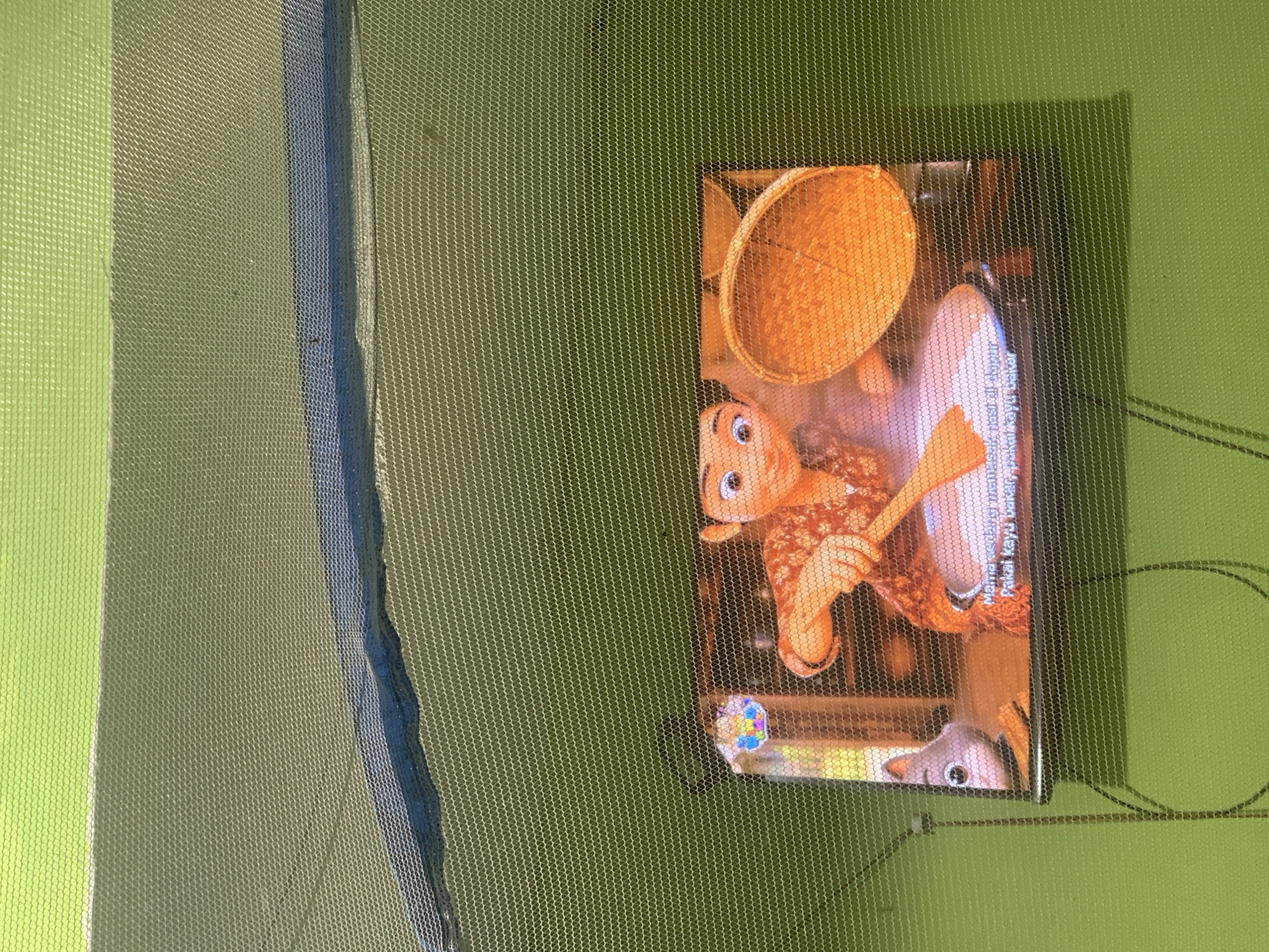An animated scene from a screen, possibly a television or tablet, is viewed through a green mesh fabric. The screen displays a young girl with dark hair, wearing a reddish-orange patterned shirt, stirring a white substance in a pan with a wooden spoon. To her left, a small grey cat-like creature peeks from behind the pan. Behind the girl, a woven basket sits on a surface. Text in Indonesian is visible at the bottom of the screen. The mesh fabric distorts the image, creating a pixelated effect. The background of the screen appears to be a kitchen interior. The overall scene is captured from a slightly upward angle, with the green mesh fabric dominating the foreground and upper portion of the image. The location in Palembang, Indonesia, is not determinable from the visual information provided.