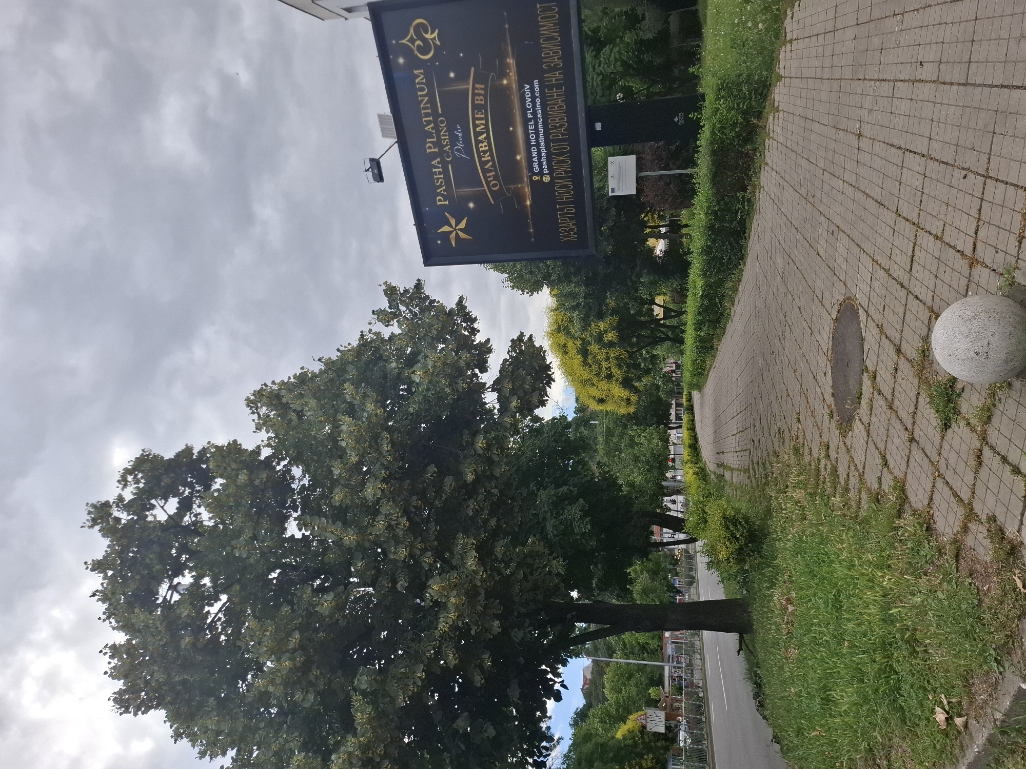 A street scene with a sidewalk and a large billboard. The billboard is dark with gold lettering and graphics, advertising "Pasha Platinum Casino." Text on the billboard is in Bulgarian and includes "ОЧАКВАМЕ ВИ" which translates to "We are waiting for you." Below that, it lists "GRAND HOTEL PLOVDIV" and a website address. At the bottom, it states in Bulgarian, "GAMBLING CARRIES THE RISK OF ADDICTION." The billboard is mounted on a tall black pole.
To the left of the billboard is a large, mature tree with dense green foliage. Further left is a road with a sidewalk and a metal fence. A few vehicles are visible on the road, and some buildings and trees line the opposite side of the street.
The sidewalk is paved with rectangular gray tiles and is bordered by grass and bushes. A spherical concrete bollard sits on the sidewalk near the foreground. The sky is overcast with gray clouds. The environment appears to be an urban or suburban setting.