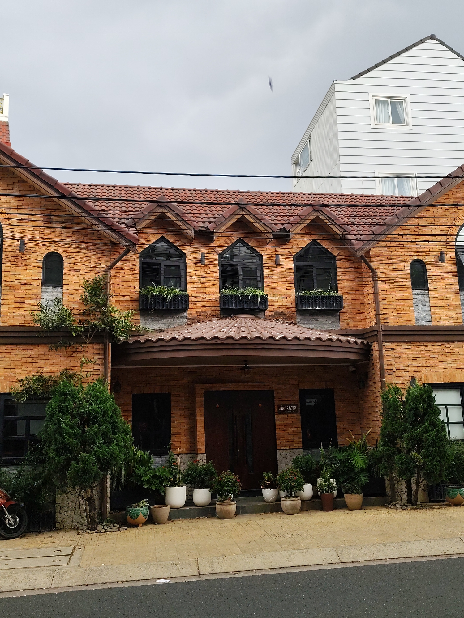 The image shows a two-story brick building with a distinctive red-tiled roof located in Da Lat, Vietnam. The facade features three arched windows on the second floor, each adorned with window boxes containing green plants. A covered porch extends over the entrance, which consists of dark wood double doors. Potted plants line the front of the building, adding to the welcoming aesthetic. Flanking the building on either side are small evergreen trees. A modern white building is visible in the background. A section of road and sidewalk are in the foreground. A motorcycle is parked near the tree on the left side. Power lines are visible above the building. The sky is overcast. A sign above the door says "Giang's House."