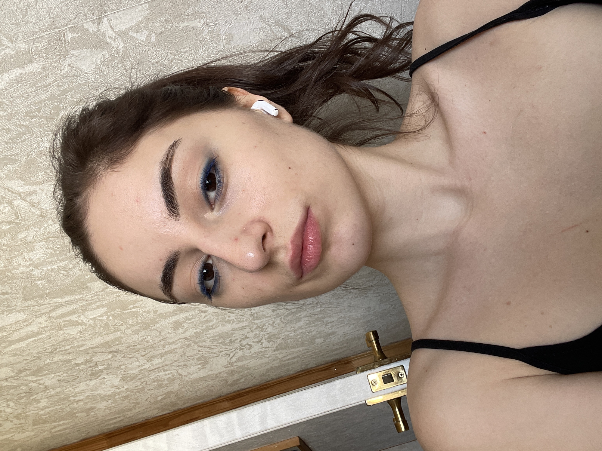 A young woman with dark hair, wearing blue eye makeup and a black tank top, poses for a selfie. She is wearing a white wireless earbud. The background includes a patterned wall and a door handle. There is no indication of the location being Moscow, Russia.