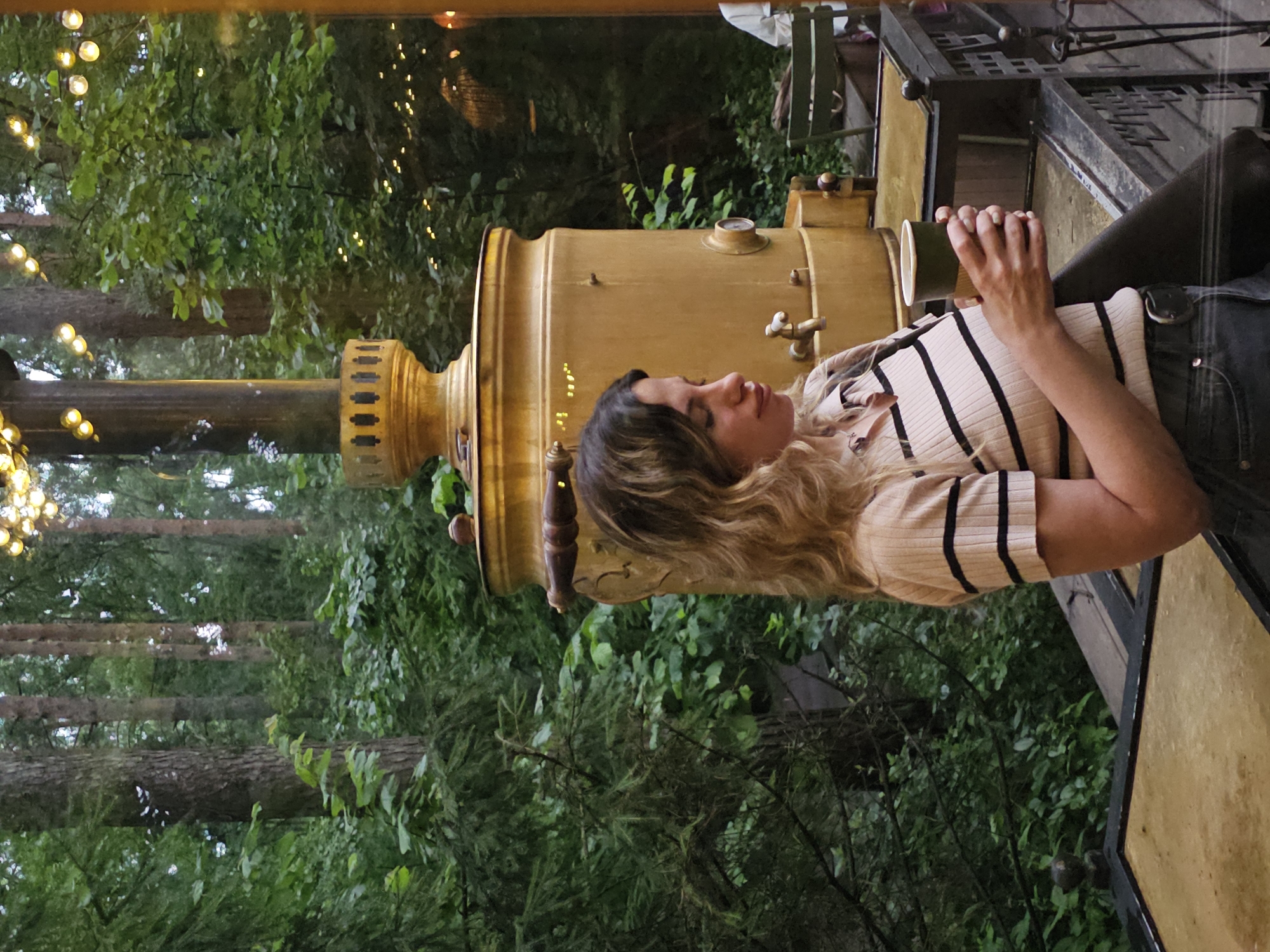 A woman with blonde, wavy hair stands in an outdoor setting, holding a dark green cup in her right hand. She is wearing a light-colored, short-sleeved shirt with horizontal black stripes and dark jeans with a belt. Behind her, to the left, is a large, golden samovar with decorative elements. The background consists of lush green trees and foliage, illuminated by a string of warm lights overhead, suggesting an evening or twilight setting. The environment appears to be a park or garden area, possibly at a cafe or restaurant, given the presence of outdoor seating and a serving counter or table in the foreground. The overall atmosphere is relaxed and serene, with the natural elements and soft lighting contributing to a tranquil mood. While the image does not provide specific visual cues to definitively place the location in Nemchinovo, Russia, it is consistent with an outdoor recreational or dining space in a wooded area.