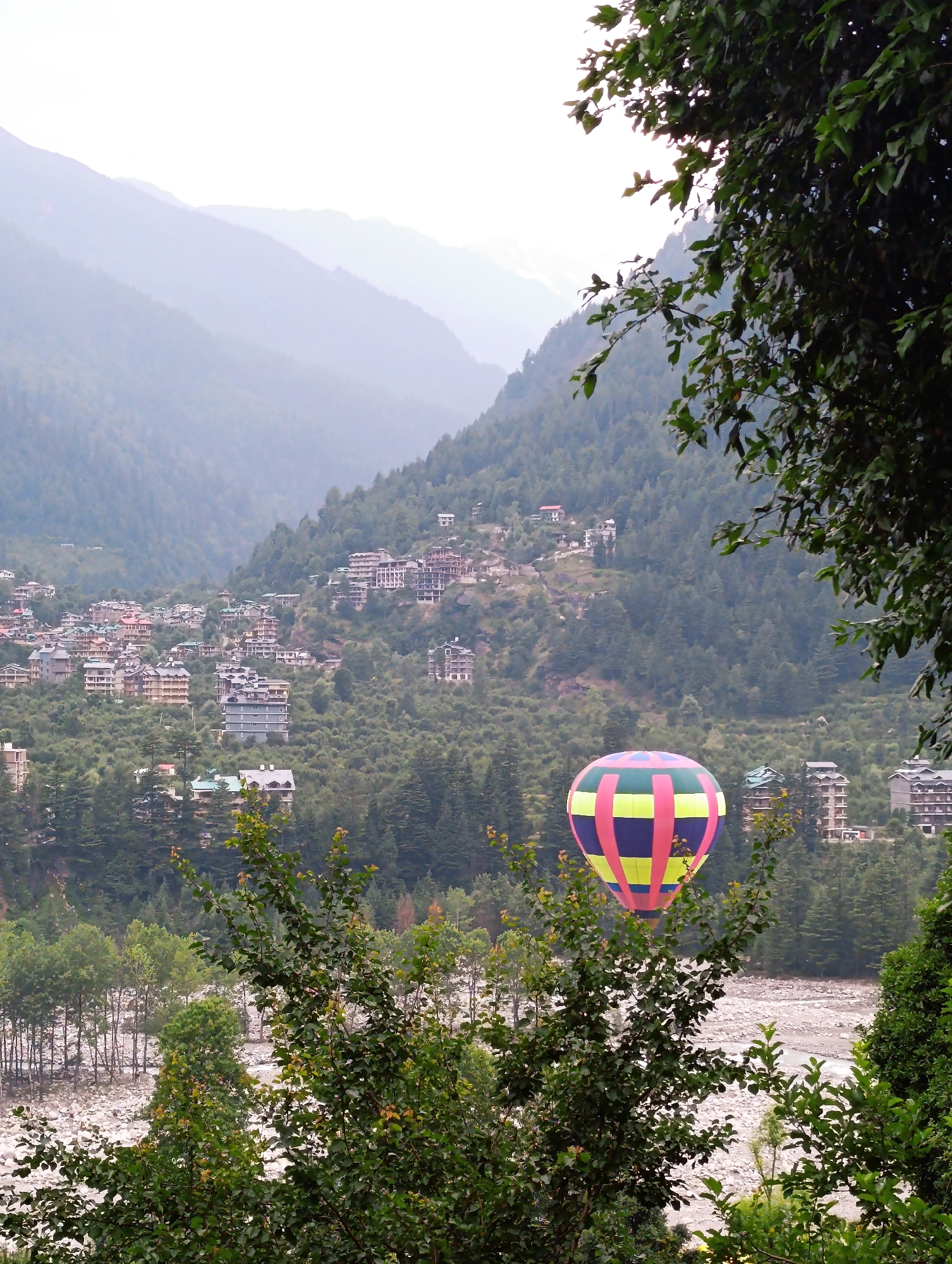 This media file depicts a serene mountain landscape in Vashisht, India. The scene is dominated by lush green mountains and a village nestled on their slopes. Buildings, varying in size and architecture, are scattered across the hillside, giving the impression of a well-established community. A hot air balloon, vibrantly colored with pink, yellow, and blue sections, floats gently in the middle ground, adding a touch of whimsical adventure to the otherwise tranquil setting.
The foreground is framed by dense foliage, with trees and branches obscuring parts of the view and adding depth to the composition. In the background, several layers of mountains recede into the distance, their forms softened by a hazy, diffused light. This suggests it could be early morning or late afternoon, or perhaps the weather is overcast. The overall atmosphere is peaceful and idyllic, with a hint of excitement from the presence of the hot air balloon. The riverbed visible at the bottom of the frame, filled with pebbles, further enhances the natural beauty of the location. No visible text is present in the image.