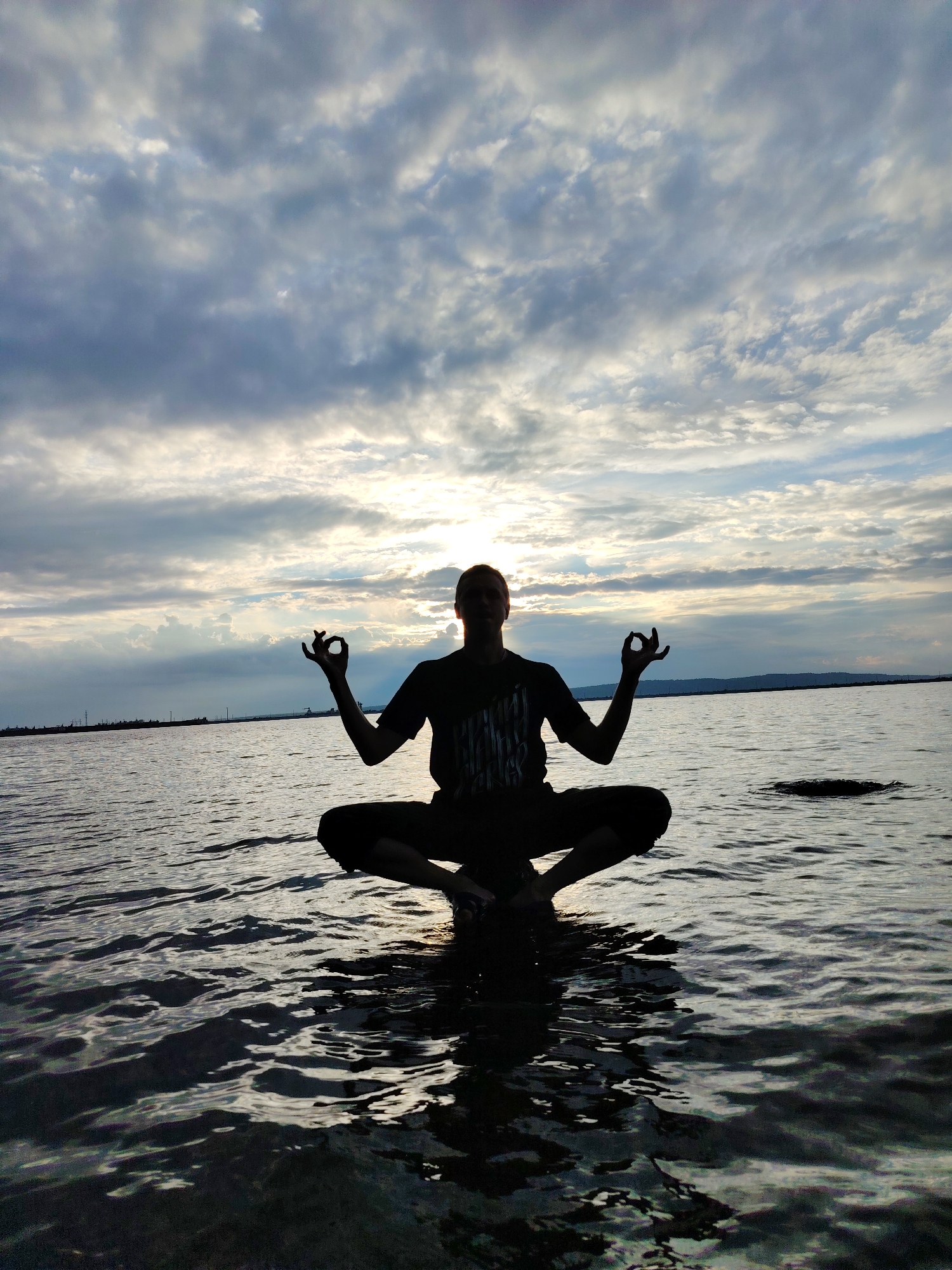 A silhouette of a person in a meditative pose sits on the surface of a body of water, appearing to float. The person is in a lotus position, with their hands resting on their knees, fingers forming the Gyan Mudra. They are silhouetted against a dramatic sky filled with clouds, illuminated by the setting sun which peeks through a break in the clouds. The water is calm, with gentle ripples reflecting the sky's light. In the distance, a hazy shoreline is visible, lined with what appear to be industrial structures or a port. The overall mood is serene and contemplative, suggesting a moment of peace and connection with nature at dusk. The setting appears to be a large body of water, possibly a lake or a river, near a populated or industrial area, given the distant structures. The time of day is clearly sunset, indicated by the low angle of the sun and the dramatic cloud formations.