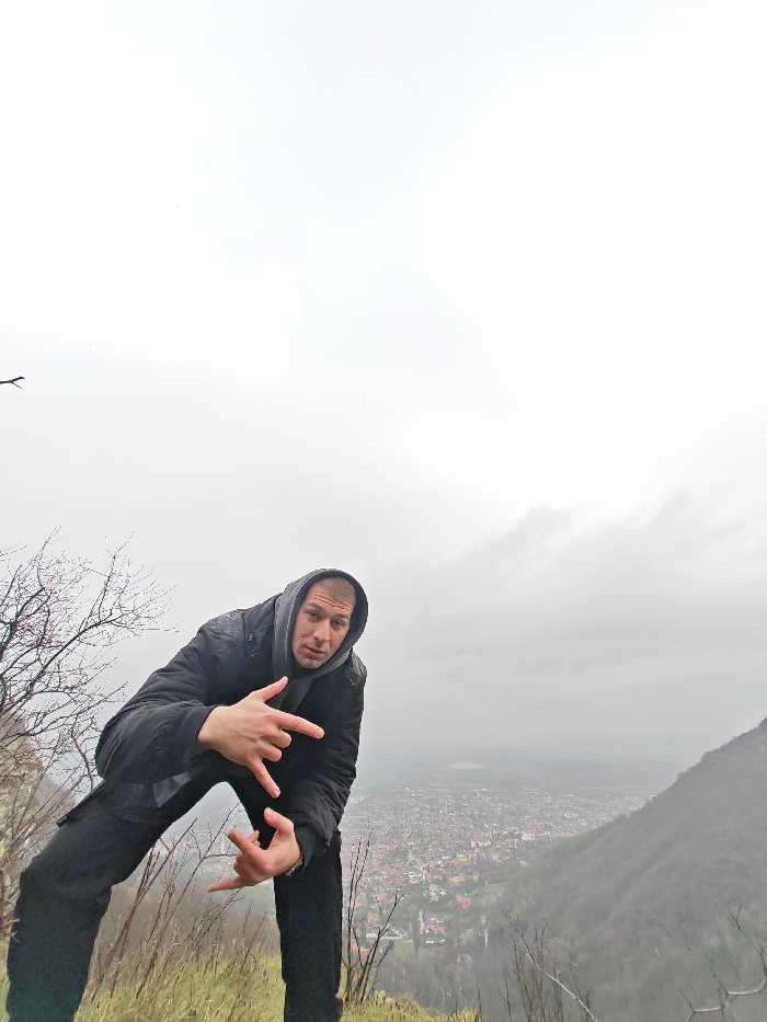 An adult male stands on a grassy hillside overlooking the city of Shumen, Bulgaria. He is positioned on an elevated natural slope, looking down upon the urban area. The male wears a dark jacket, a grey hooded sweatshirt with the hood raised, and dark pants. His body is leaned forward at the waist with slightly bent knees, and he faces the camera directly. Both hands are raised to chest height, forming a gesture with the index and pinky fingers extended. To the left of the subject, bare tree branches are visible. The city of Shumen is discernible in the mid-ground, characterized by densely packed buildings, many with red-tiled roofs, extending into the distance. A body of water is faintly visible beyond the city horizon. A dark, forested mountain slope occupies the far right of the frame. The sky is overcast and appears bright white, dominating the upper portion of the image.