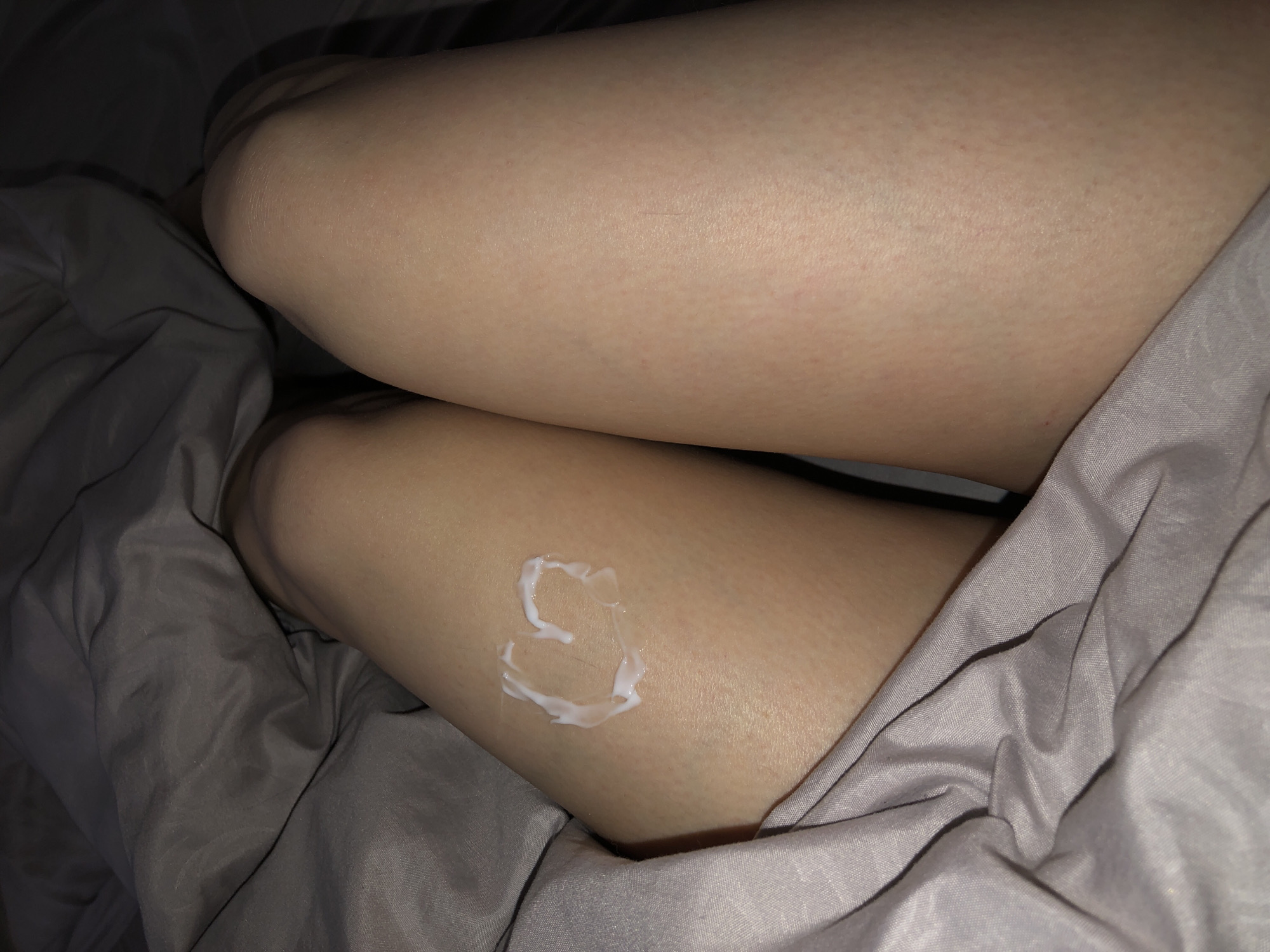 The image shows a close-up of a person's thighs resting on a gray bedsheet. On the lower thigh is a heart shape formed from a white substance, possibly lotion. The lighting is dim, and the focus is sharpest on the heart shape and the texture of the skin. The location is not identifiable from the image.