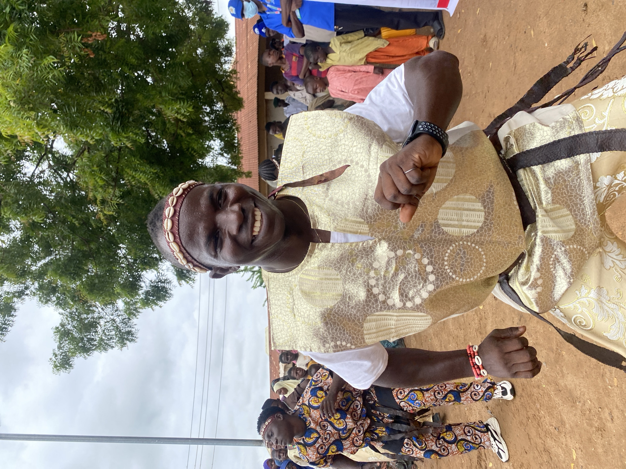 In Rigachikun, Nigeria, a man is captured mid-dance, adorned in a gold vest with intricate patterns, a white t-shirt, and ornate gold pants with similar detailing. He wears a headband with shells and a wristband of red beads and shells. The man is smiling broadly, his right arm extended as if in a dynamic pose. Behind him, a crowd of people, including men, women, and children, observe. One person in the background wears a blue shirt and a face mask. The setting appears to be an outdoor gathering, with a dirt ground and a tree with green foliage overhead, suggesting a warm climate. The sky is overcast, with visible power lines and a building with a red roof in the distance.