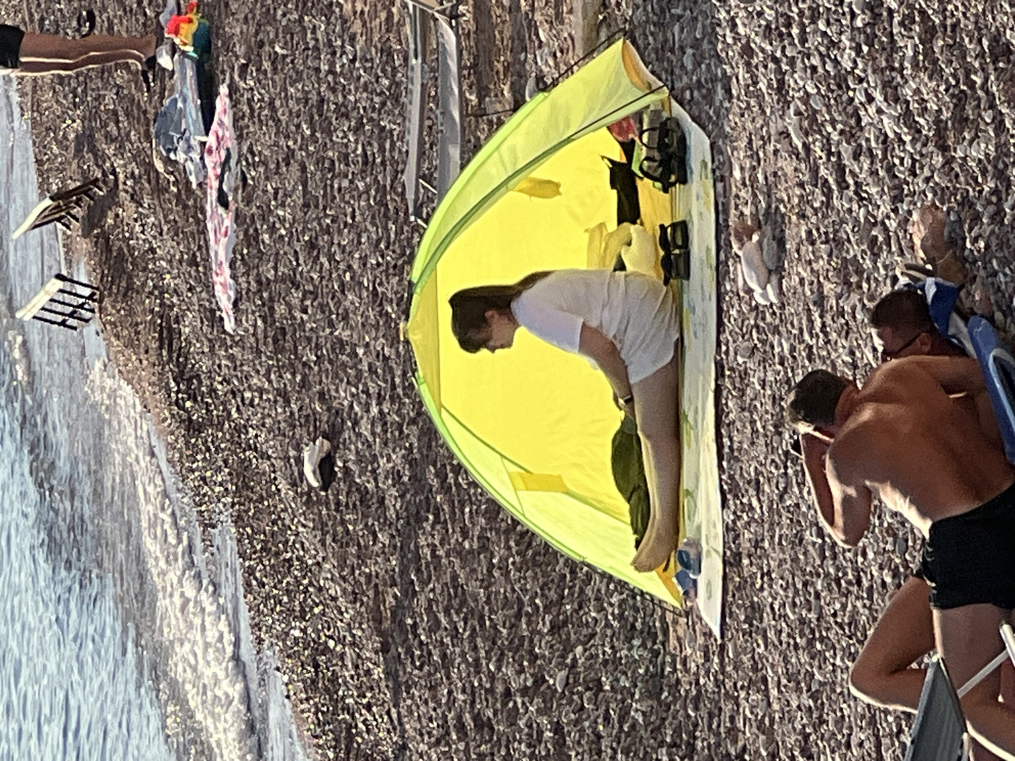 In Sveti Stefan, Montenegro, on a pebble beach, a woman sits inside a bright yellow tent. She is facing left, wearing a white t-shirt and white shorts. Her legs are stretched out in front of her. A green mat is laid out under the tent, and a pair of blue sandals and two pairs of black sandals are on the mat. Behind the tent, in the foreground, two men are lying on loungers. One man, in the lower left of the image, is lying on his back, wearing black swim trunks. The other man is lying on his front, his back to the camera, wearing black swim trunks. The sea is visible in the background, with gentle waves lapping the shore. Several beach chairs are also visible further up the beach.