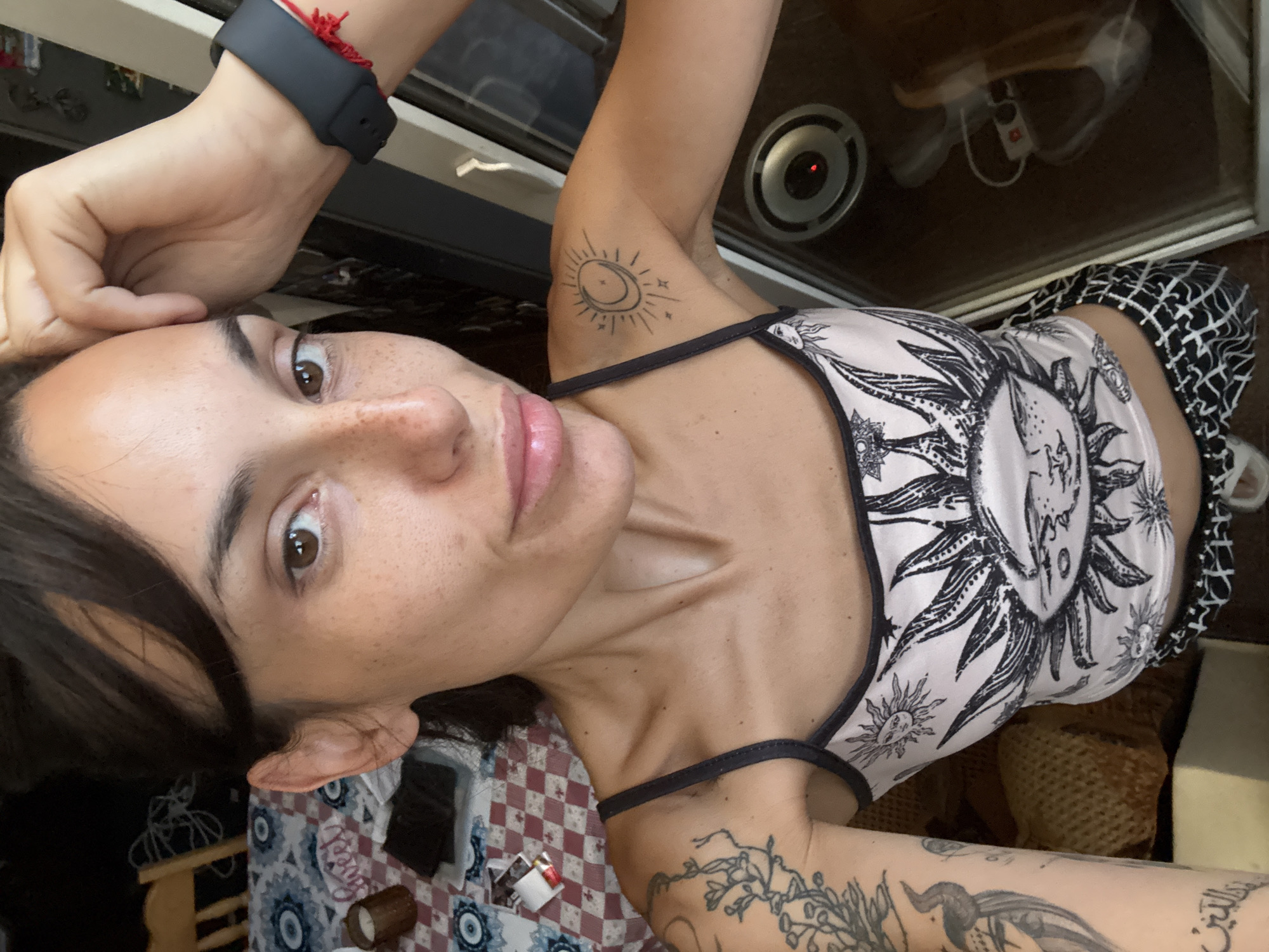 The image shows a close-up, top-down view of a person lying down. The person has dark hair, brown eyes, and visible freckles on their face. They are wearing a tank top with a black and white sun and celestial pattern, and patterned shorts. There are several tattoos visible on their arms and shoulder, including a sun and moon design on the upper arm and floral designs on the forearm. On their left wrist, they are wearing a black smartwatch with a red string tied to it.
The setting appears to be indoors, possibly a bedroom or living area. There is a patterned tablecloth or blanket visible beneath the person, with red and white checkered elements and blue evil eye motifs. In the background, parts of furniture, including a wooden chair and what looks like a dresser or cabinet, are visible. Behind the person's head, a circular object with a red light in the center, possibly a speaker or light fixture, is visible, reflecting in a glass surface. A power outlet with a cord plugged in is also visible in the reflection.
The lighting suggests it is daytime. The person's expression is neutral, with their eyes looking towards the camera. The overall mood is casual and relaxed. No discernible text is visible in the image.