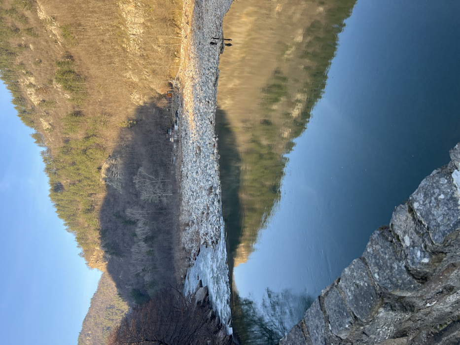 The scene depicts a river valley in the vicinity of Diadovtsi, Bulgaria, observed from an elevated stone structure, likely a bridge. A stone railing occupies the lower left foreground. The midground features a river with a calm, reflective surface in the section closest to the viewer, mirroring the opposing sunlit mountain slope and sky. Further upstream to the left, the river exhibits a turbulent, flowing current. The right bank of the river consists of a wide expanse of light-colored, rounded rocks. Two human figures are standing upright on this rocky bank at the water's edge on the right side of the image, their reflections visible in the still water. Beyond the river, steep mountain slopes rise on both sides. The right mountain slope is illuminated by direct sunlight, revealing a mix of evergreen coniferous trees and leafless deciduous trees, interspersed with some exposed rock faces. Several small buildings or structures with dark roofs are situated along this sunlit riverbank. The left mountain slope is largely in shadow, also covered with a mix of evergreen and bare deciduous trees. A clear blue sky is visible above the mountain peaks. The bare deciduous trees suggest a cooler season.