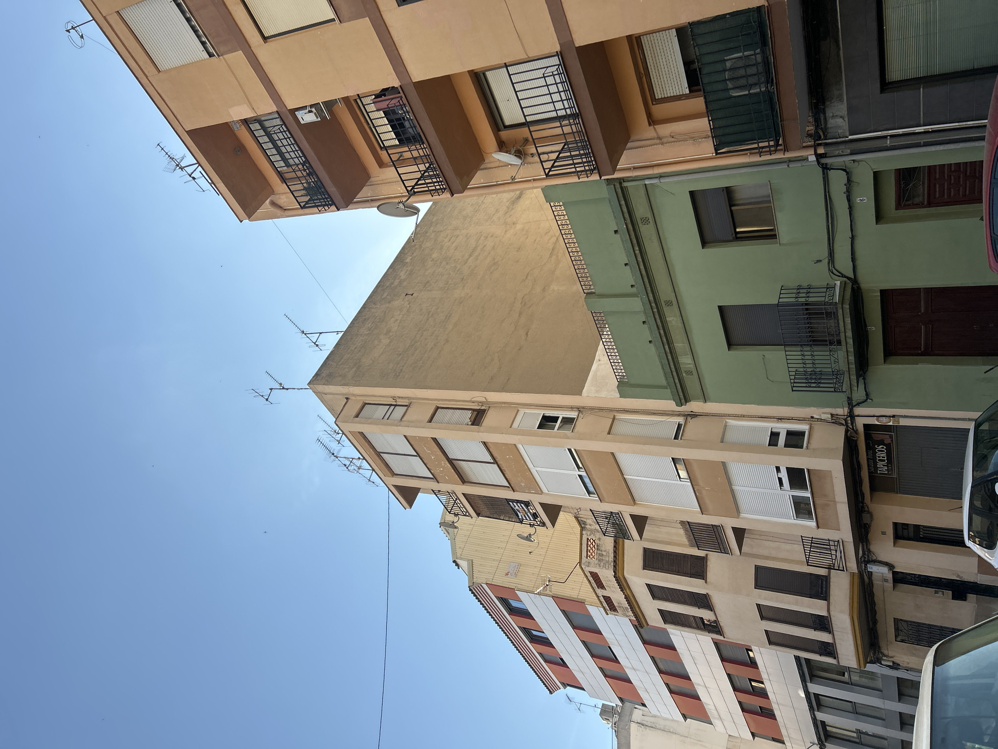This is a low-angle, outdoor shot of buildings in Castelló De La Plana, Spain, under a clear blue sky. The sun appears to be high in the sky, suggesting it is daytime. The buildings are of various heights and colors, including beige, light green, and some with red and white striped facades. Several windows with closed blinds and some with wrought-iron balconies are visible. Antennas are mounted on the rooftops of some buildings. There is a sign on one building that reads "TAPICEROS." The perspective suggests a narrow street or alleyway between the buildings. No people or specific activities are depicted.