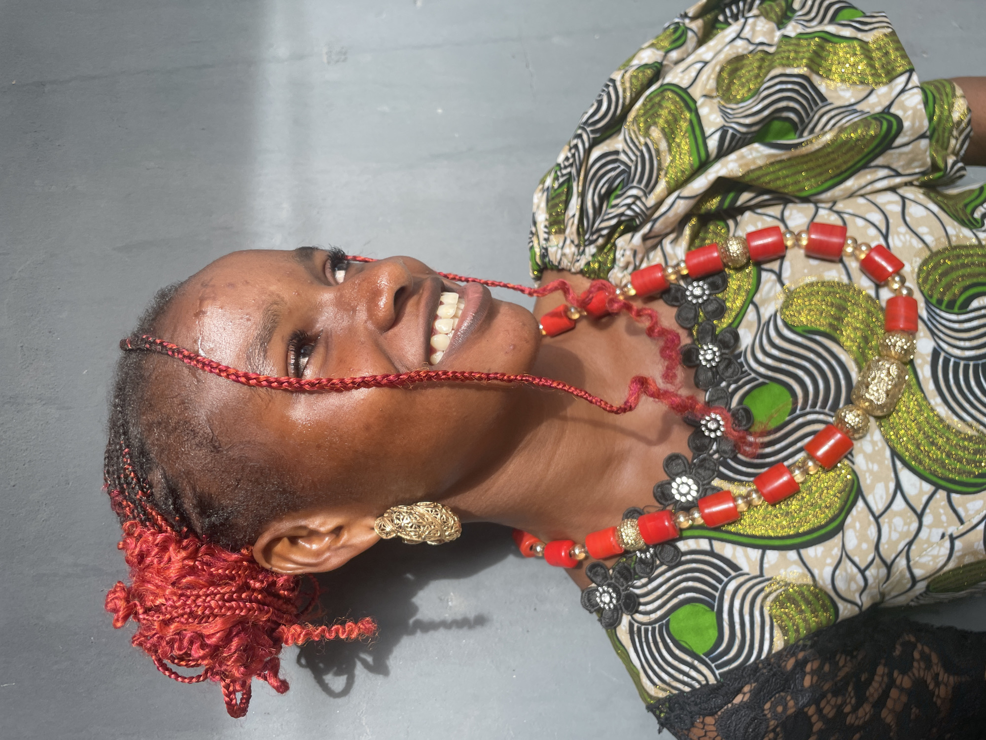 A joyful young woman with dark skin and distinctive red braided hair is captured in a close-up, horizontal shot. She is pictured lying down on a light grey surface, looking upwards with a broad, radiant smile that reveals her white teeth. Her head is tilted slightly, and a subtle sheen, possibly sweat, is visible on her forehead and cheeks. Her dark eyes are directed towards the top left of the frame. Her red braids are styled in cornrows, gathered into a bun-like cluster on the left side of her head.
She is dressed in vibrant attire, featuring an Ankara-style fabric with a rich pattern of green, black, white, and beige. The green sections of the fabric appear to have a glittery texture. A hint of black lace trim is visible on her sleeve. Her accessories are prominent: a multi-strand necklace adorned with striking red coral-like cylindrical beads interspersed with gold-colored spherical beads, and accented with black floral embellishments that have white or silver centers. A intricate, gold-colored filigree earring is visible on her right ear.
The background consists of a plain, light grey surface, which could be concrete or tiles, showing some faint texture. The lighting is bright and appears natural, suggesting a daytime scene in an outdoor or brightly lit indoor environment. The woman's expression conveys happiness and contentment. The traditional attire and coral bead jewelry are indicative of cultural styles found in West Africa, aligning with the provided context of Badagry, Nigeria. There is no visible text in the image.