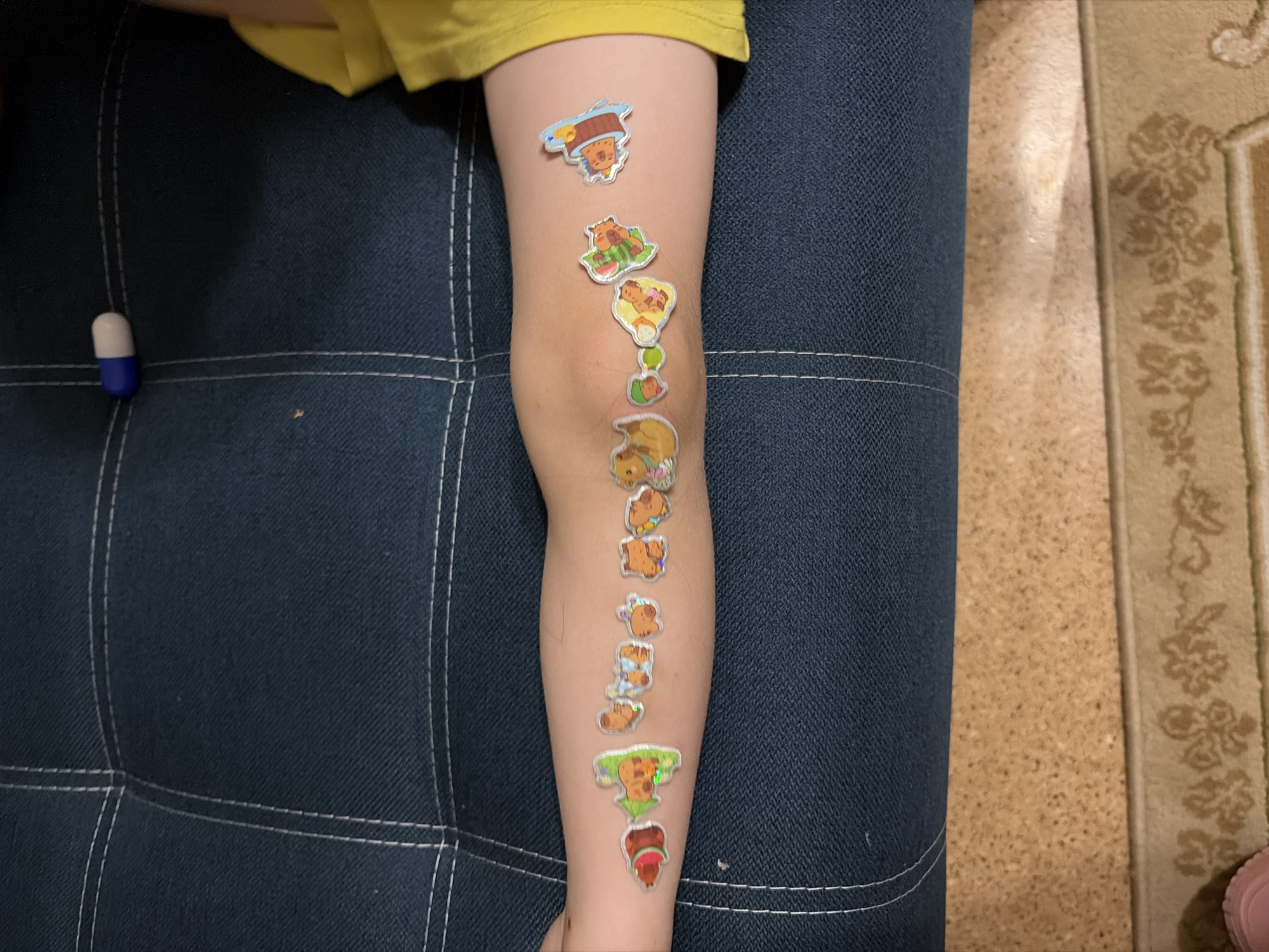 An image depicts a close-up view of a person's arm with several stickers applied to it. The stickers are small, colorful, and feature cartoonish animal designs, resembling characters like bears and ducks. They are arranged in a line along the arm, some appearing to be in a sequential or narrative arrangement. The arm itself appears to be that of a child or young person, with light skin and visible fine hairs. The arm is resting on a dark blue, quilted surface, likely upholstery. In the background, a yellow fabric is visible, possibly clothing. There is also a blue and white pill-shaped object positioned on the quilted surface near the top edge of the frame. The overall setting appears to be indoors. There is no specific information about the location in Tambov, Russia.