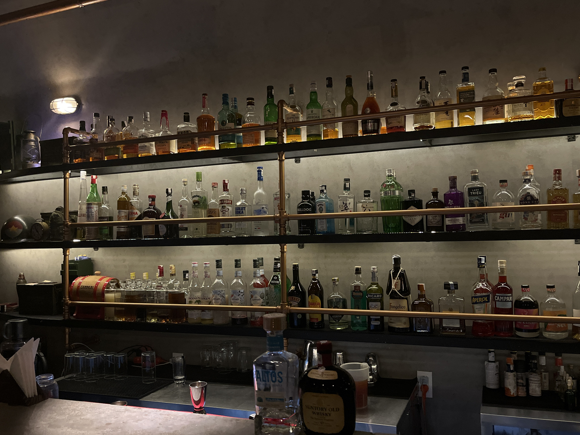 This image shows a well-stocked bar, likely in Đà Nẵng, Vietnam, given the presence of Vietnamese brands like "LADY TRIỀU" vodka and "SỐNG CÁI" gin among the more common international brands. The bar has three tiers of shelves filled with various bottles of liquor. The top shelf features a variety of brown liquors and whiskeys. The middle shelf contains a mix of clear and colored liquors. The bottom shelf contains liqueurs and more assorted bottles. Copper-colored pipes support the shelves. In the foreground, the bar counter is visible with glassware, napkins, a shot glass, and bottles of Altos tequila and Suntory Old whisky.