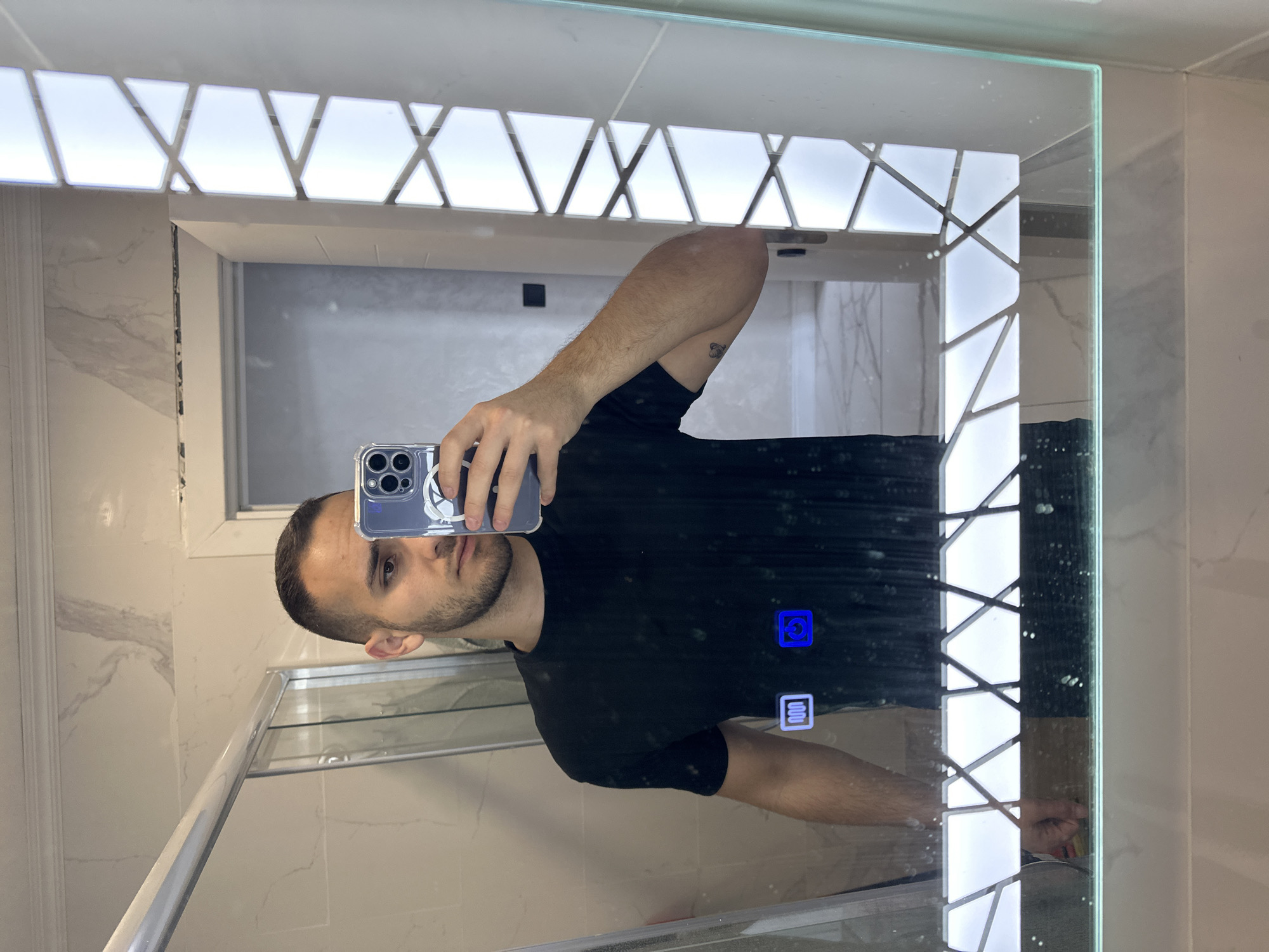 A young man with short dark hair and a beard, wearing a black t-shirt, takes a mirror selfie in a modern bathroom. He is holding an iPhone in his right hand, capturing his reflection. The mirror has integrated LED lighting around its edges, with a geometric pattern of white and translucent panels. Below the lighting, a blue illuminated icon resembling a power button is visible on the mirror's surface. To the left of the power button, another blue illuminated icon depicts wavy lines, suggesting a heating or defogging function. The bathroom features marble-patterned walls and a glass shower enclosure. The background reflected in the mirror shows a doorway and a tiled floor. The scene is set in Varna, Bulgaria, specifically within a residential bathroom.
