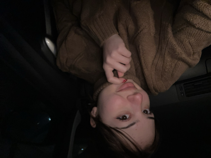 A young woman with light skin, brown eyes, and dark hair is wearing a brown, textured sweater. She’s resting her hand against her chin, and her nails are painted with small red dots. The scene is dimly lit, with the woman appearing to be inside a vehicle. The interior details include a car door, a window, and a ventilation system. The angle suggests the photo was taken upside down.