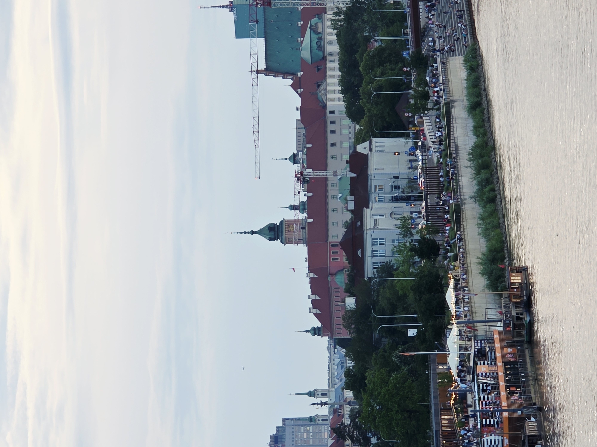 This is a wide-angle, eye-level photograph of the Vistula River embankment in Warsaw, Poland, with the Old Town and New Town skyline in the background under a pale, overcast sky. In the foreground, the rippling, brown water of the Vistula River occupies the bottom third of the frame. Moored to the embankment is a multi-level floating structure, painted orange and black, with various decks, awnings, and people visible. To the right of the floating structure, the embankment rises, lined with a concrete wall and a stepped seating area where numerous people are gathered, some sitting and others standing, enjoying the view. Behind the embankment, a lush green line of trees separates the riverfront from the city buildings.
Dominating the mid-ground and background is a panorama of Warsaw's historic architecture. The most prominent building is the Royal Castle, characterized by its red-tiled roofs and the green dome of its clock tower. Adjacent to it, other historic buildings with red roofs and lighter facades are visible, including what appears to be St. John's Archcathedral with its pointed spire. Further to the left, modern high-rise buildings, including a glass skyscraper, punctuate the skyline, alongside older, spired buildings, one of which is a tall, dark obelisk. Several construction cranes are positioned throughout the cityscape, indicating ongoing development. The overall scene depicts a vibrant urban environment with a mix of historical grandeur and modern activity, set against a soft, diffused light.