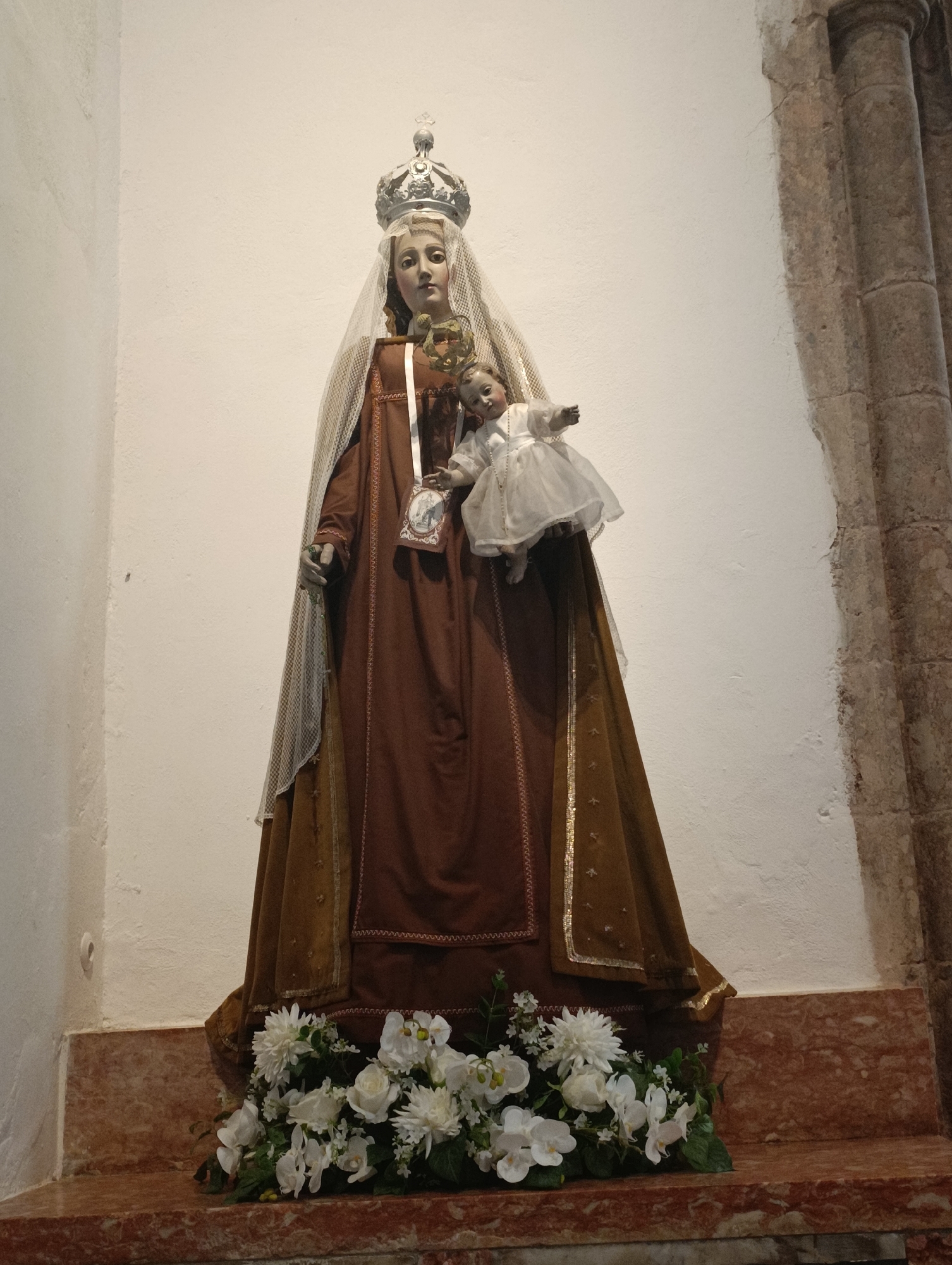 The image shows a statue of the Virgin Mary holding the infant Jesus. The statue is adorned with a crown and veil, and the Virgin Mary is wearing a brown robe. The infant Jesus is dressed in a white gown. The statue is placed on a marble altar, which is decorated with a bouquet of white flowers. The background of the image is a plain white wall, with a stone column on the right side. This statue is located in the Capela de Nossa Senhora da Conceição da Pena, a chapel in the historic center of Setúbal, Portugal, near the Praça de Bocage.