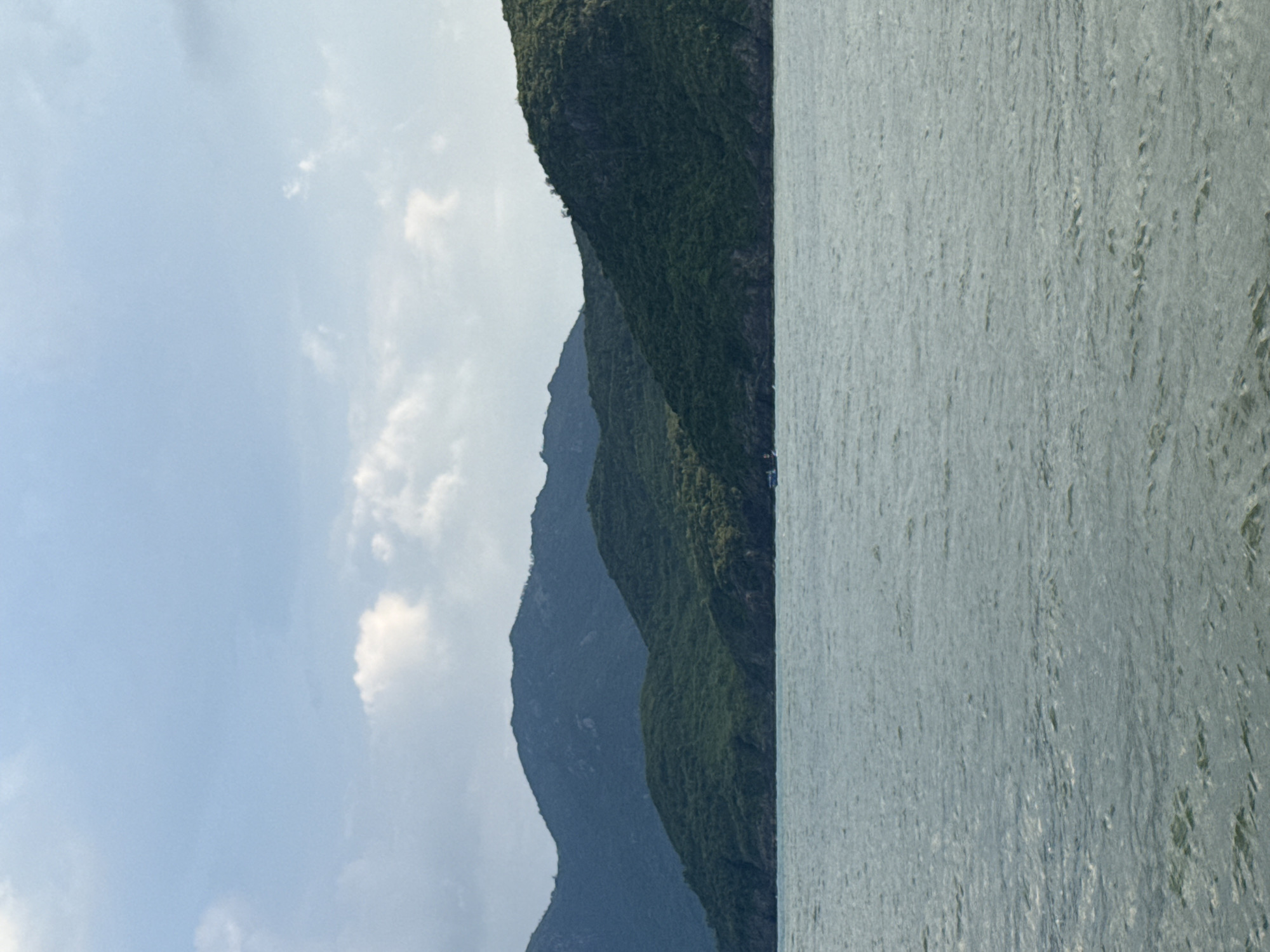 The image depicts a serene coastal landscape with rolling green hills meeting the sea under a partly cloudy sky. The water in the foreground is a tranquil blue-grey, with gentle ripples suggesting a calm day. In the middle ground, lush, verdant hills rise steeply from the shoreline, their slopes covered in dense vegetation. The furthest hills are darker and more distant, fading into a dusky blue hue against the horizon. A small, white boat is visible near the shoreline, adding a touch of human presence to the natural scenery. The sky is a soft blue, dotted with scattered white clouds, some of which are wispy and others more substantial. The overall impression is one of natural beauty and peacefulness. This scene is typical of the coastline found in Nha Trang Ward, Vietnam, a region renowned for its picturesque bays, islands, and mountainous terrain meeting the sea.