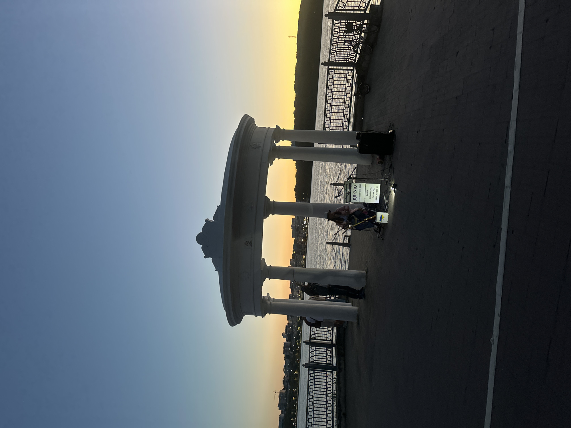 At dusk, a white rotunda with classical columns stands on a paved embankment overlooking a body of water. The sky transitions from deep blue to orange near the horizon, where a city skyline and a distant radio tower are visible. In front of the rotunda, a street performer, possibly a musician, is set up with a microphone, amplifier, and a sign that reads "ДЯКУЮ" (Thank You) in Ukrainian, along with a phone number and the phrase "ВАША УЛЮБЛЕНА ПІСНЯ НА ЗАМОВЛЕННЯ" (Your favorite song by order). A bucket with a Ukrainian flag is also present. Two individuals are standing near the rotunda's columns, and two bicycles are parked along the railing of the embankment. The scene depicts a public space in Ternopil, Ukraine, during an evening performance.