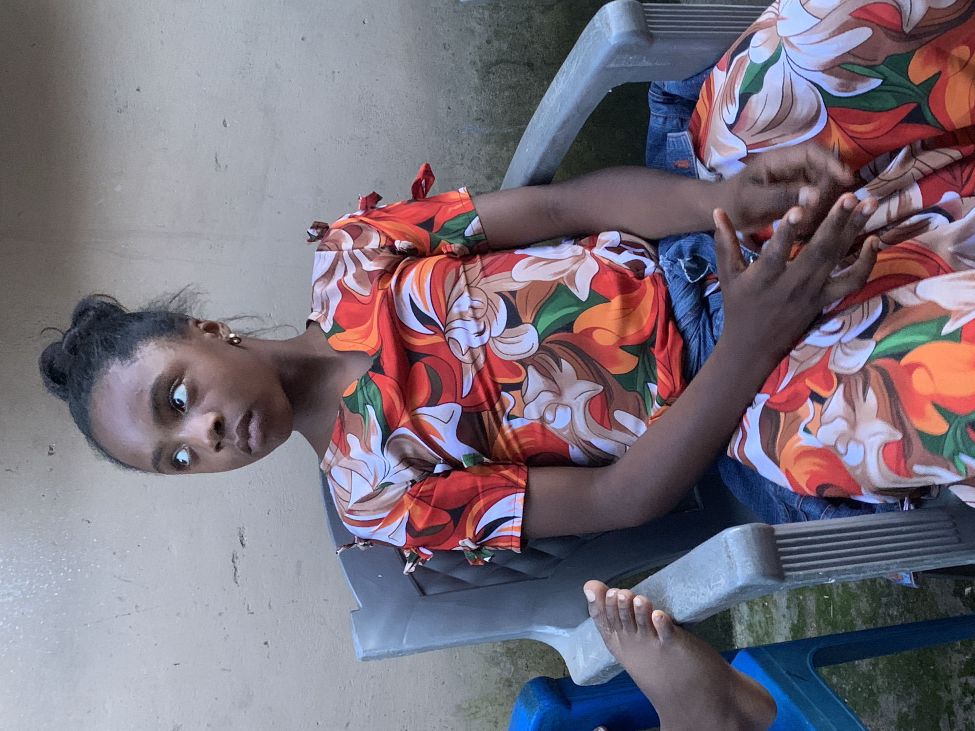In Jos, Nigeria, a young Black girl is seated in a gray plastic chair outdoors. She is looking upwards and to her left, her hair styled in a bun. She wears a colorful floral-patterned dress with red, orange, white, and green tones. Her hands are clasped in her lap. To her left, the foot of another person is visible, resting on the armrest of a blue plastic chair. The background consists of a textured, light-colored wall.