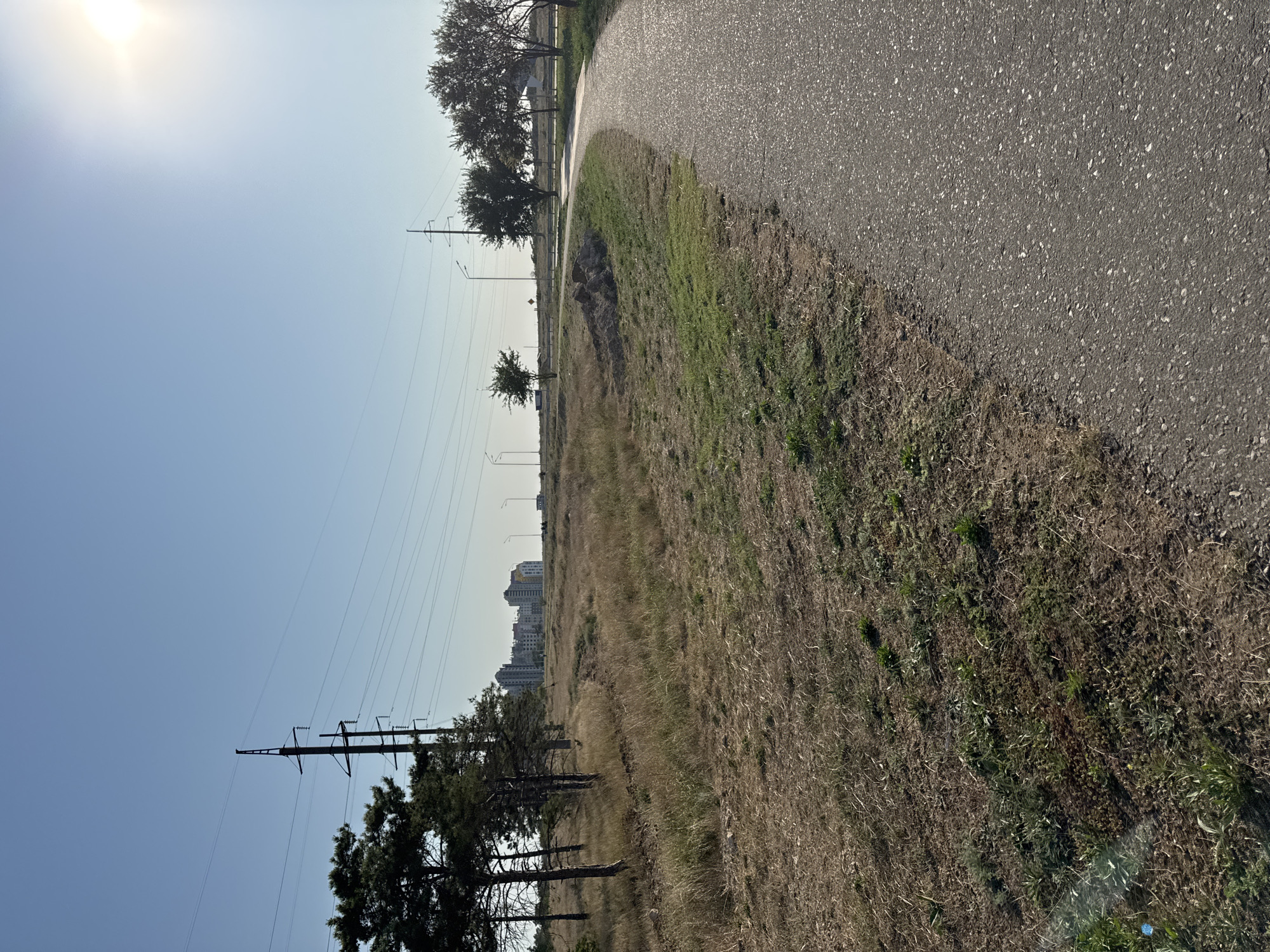 This image depicts a paved pathway curving to the right, bordering a dry, grassy field. In the distance, several tall buildings are visible, characteristic of urban development. Power lines and utility poles stretch across the scene, indicating infrastructure. The sky is clear and bright, with the sun shining from the upper right, casting a slight glare. The overall setting appears to be an outdoor, semi-urban or suburban environment, likely in Yevpatoriya, Ukraine, given the context. There are no people or animals visible in the image.