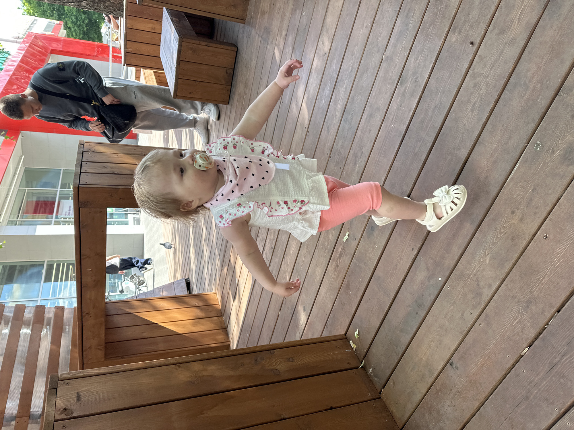 A sunny outdoor scene captures a young toddler standing on a wooden deck, part of an urban public space in Nizhny Novgorod, Russia. The toddler, with blonde hair, wears a white patterned top, a pink bib with heart motifs, pink leggings, and white sandals, and has a pacifier in their mouth. They look towards the left of the frame with one arm slightly outstretched.
In the mid-ground, an adult male stands near a wooden bench, wearing a grey sweatshirt with a distinctive patch (likely a brand logo) on the left arm, grey sweatpants, and white sneakers. He carries a black cross-body bag and appears to be looking down. The background features modern buildings with red and white facades and large glass windows, reflecting the bright daylight. Wooden structures, including decking, benches, and barriers, define the immediate environment. Strong sunlight casts clear shadows, indicating a bright, clear day. A pigeon is visible in flight in the background. The overall setting suggests a pleasant day in a pedestrian or cafe area.