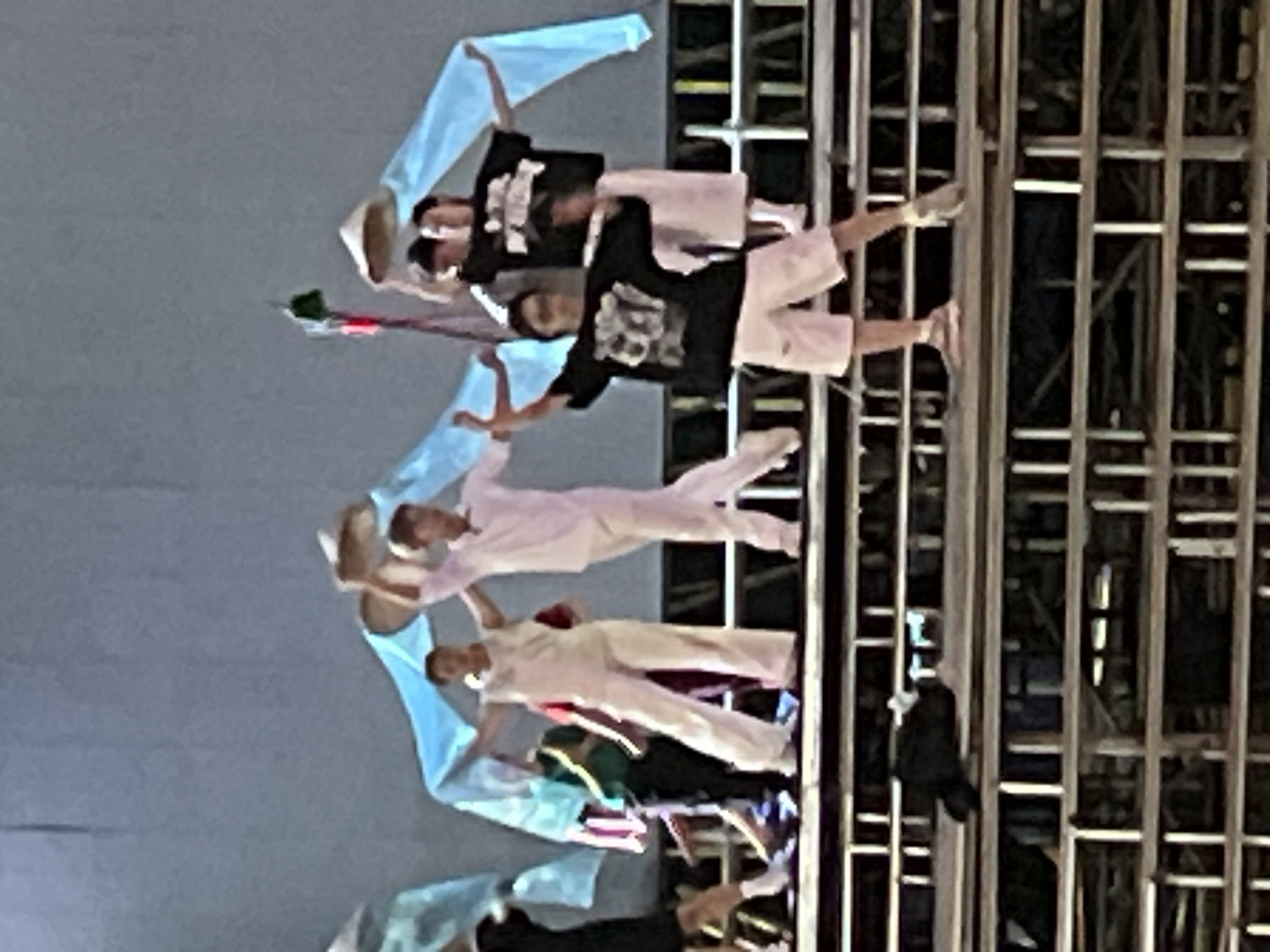 A group of dancers performs on a tiered stage. The dancers are dressed in white or black shirts and white pants or shorts. Some of the dancers are wearing conical hats. Blue fabric is draped behind the dancers and extends upwards, creating a backdrop. The stage appears to be constructed of metal scaffolding. The overall lighting suggests an outdoor evening performance. The image is slightly blurred, indicating a quick capture or low resolution. While the image itself does not provide definitive location information, this type of performance is common in public spaces and cultural events in cities like Nha Trang, Vietnam.