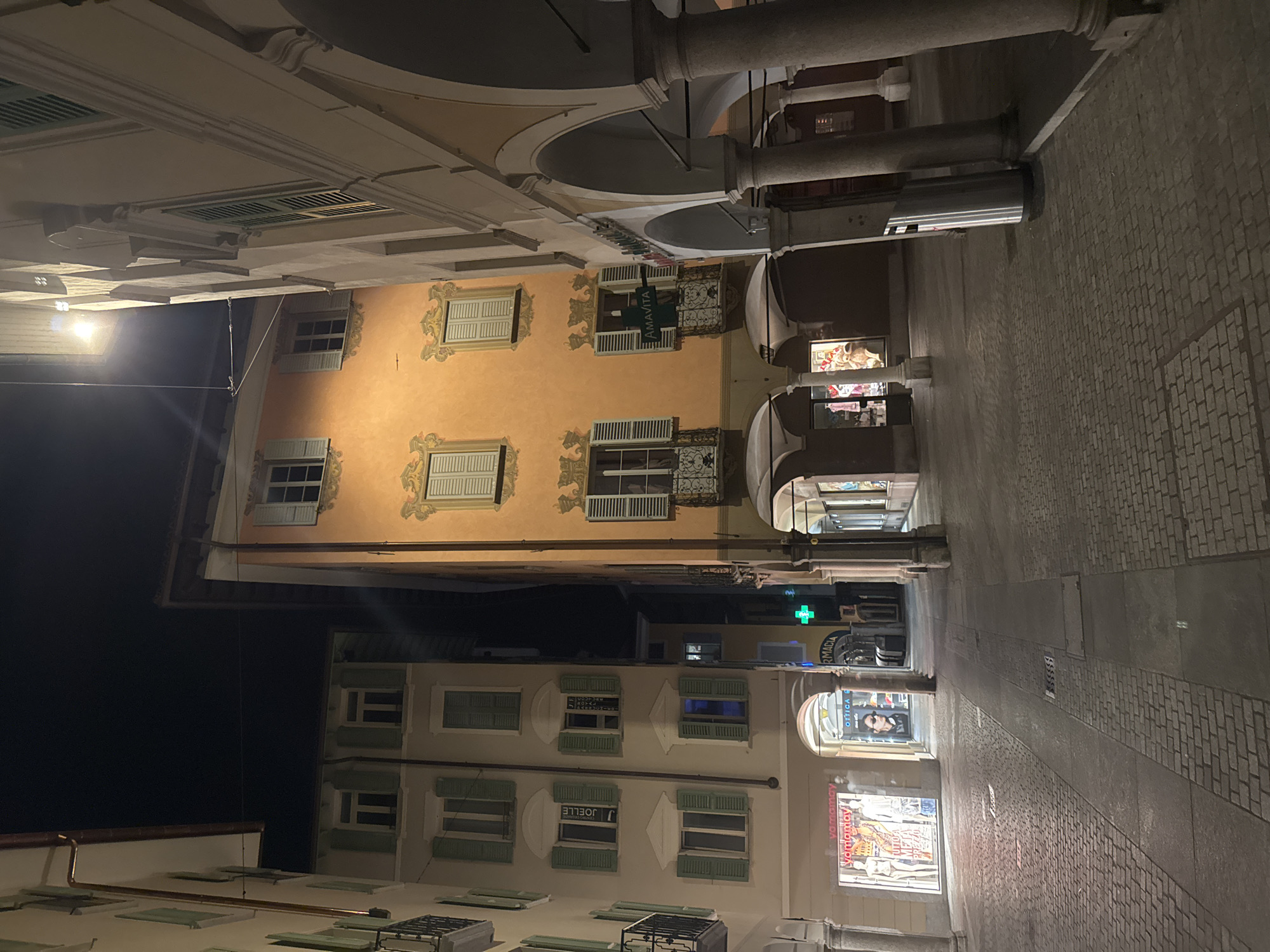 The image depicts a nighttime view of a cobblestone street in Lugano, Switzerland, lined with multi-story buildings. The street is illuminated by artificial light, creating long shadows.
The buildings on the left are lighter in color and feature green shutters on their windows. The building on the right is a prominent orange structure with decorative elements around its windows. Porticoes with columns line the right side of the street, creating an arcade.
Several shops are visible on the ground floor, including one with mannequins displaying clothing, a shop labeled "OTTICA," and a pharmacy with a green cross sign. A trash receptacle is positioned near the columns on the right. The sky is dark, indicating it is nighttime. There are no people visible in the scene.