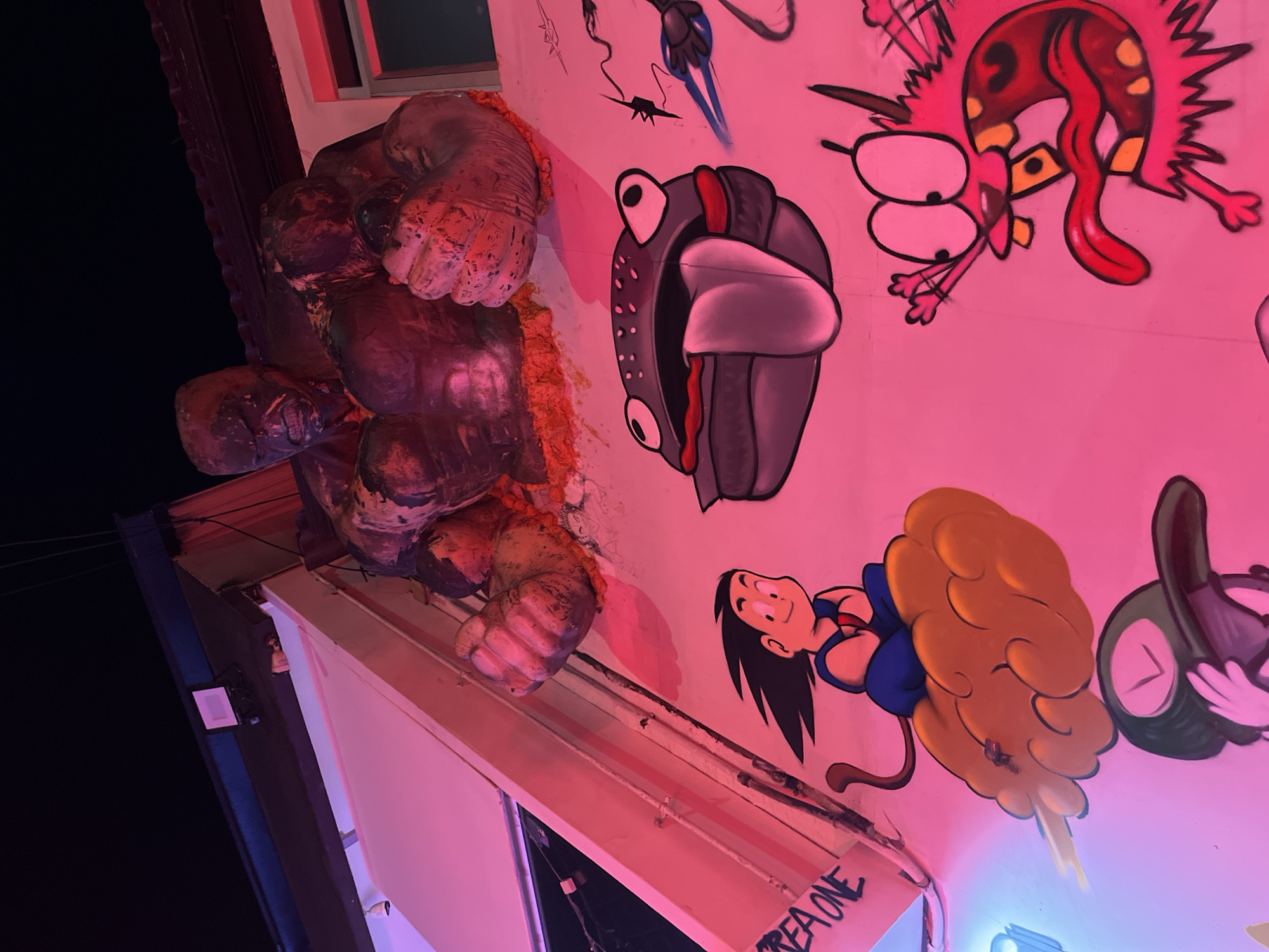 This is a low-angle shot of a building exterior at night. On the wall is a 3D sculpture resembling a monster's hand bursting through the wall, followed by graffiti-style artwork including cartoon characters. The name "REA ONE" is visible on the bottom portion of the wall, suggesting it is located in an area where graffiti is prominent. The location is unknown.