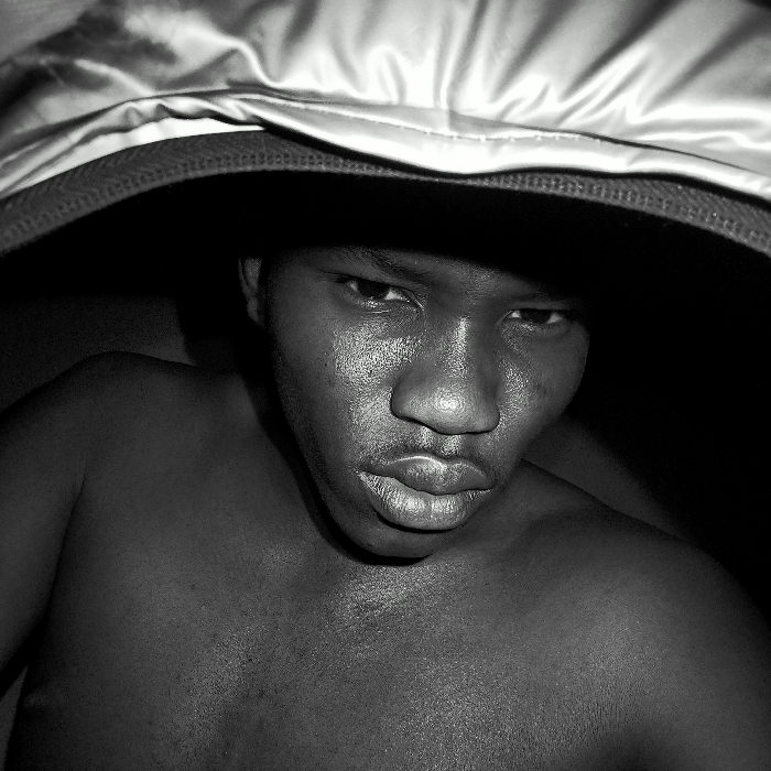 A striking black and white close-up features a shirtless man looking directly at the viewer. Part of a hood is pulled over his head. His face is illuminated, showcasing distinct features like his eyes, nose, and lips. Skin texture is visible, adding depth to the image. The lighting casts shadows, emphasizing the contours of his face and upper body. The overall composition has a raw and intense feel.
