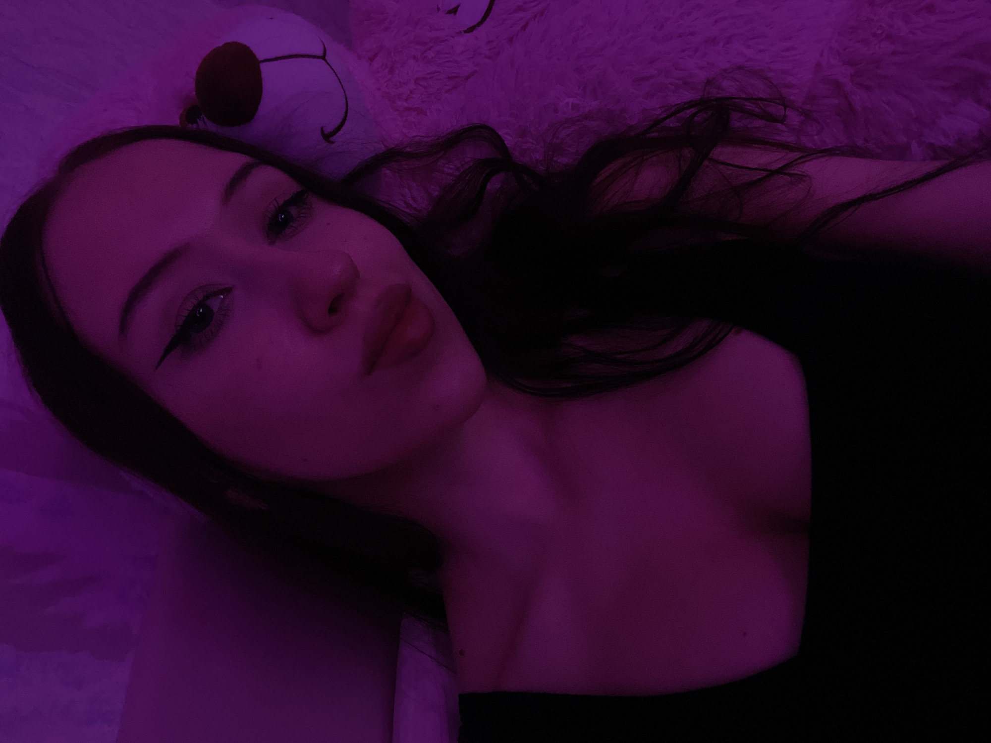This is a selfie of a young woman with long, dark hair, taken indoors under purple lighting. She is wearing a black top. To her right, a large, fluffy white stuffed bear is visible, with a brown nose and stitched facial features. The background appears to be a wall or draped fabric, also illuminated in purple. The overall ambiance is intimate and soft due to the lighting and the presence of the stuffed animal. There is no indication of the location being Votkinsk, Russia.