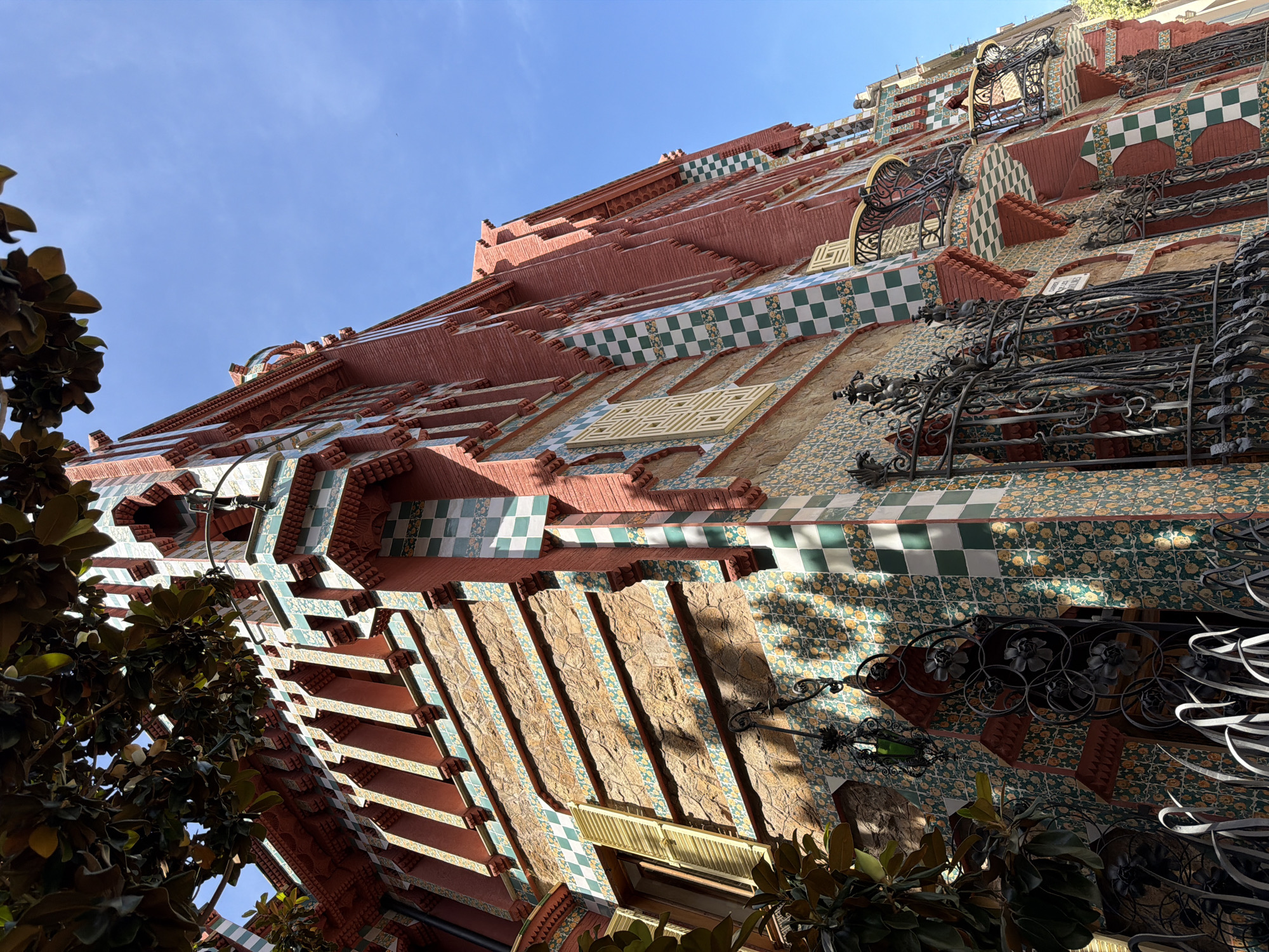 The image shows a low-angle shot of the Casa Vicens, located in Barcelona, Spain. The building's facade is richly decorated with colorful ceramic tiles, featuring a mosaic of green and white checkerboard patterns, as well as floral motifs in shades of yellow and green. The red brickwork is visible in sections, and the building has ornate architectural details, including arched windows, decorative cornices, and intricate wrought-iron balconies. A large tree with dark green leaves is visible in the upper left corner, casting shadows on the building. The sky above is a clear, bright blue. The scene appears to be bathed in sunlight, highlighting the vibrant colors and textures of the architecture. There are no people visible in the frame.