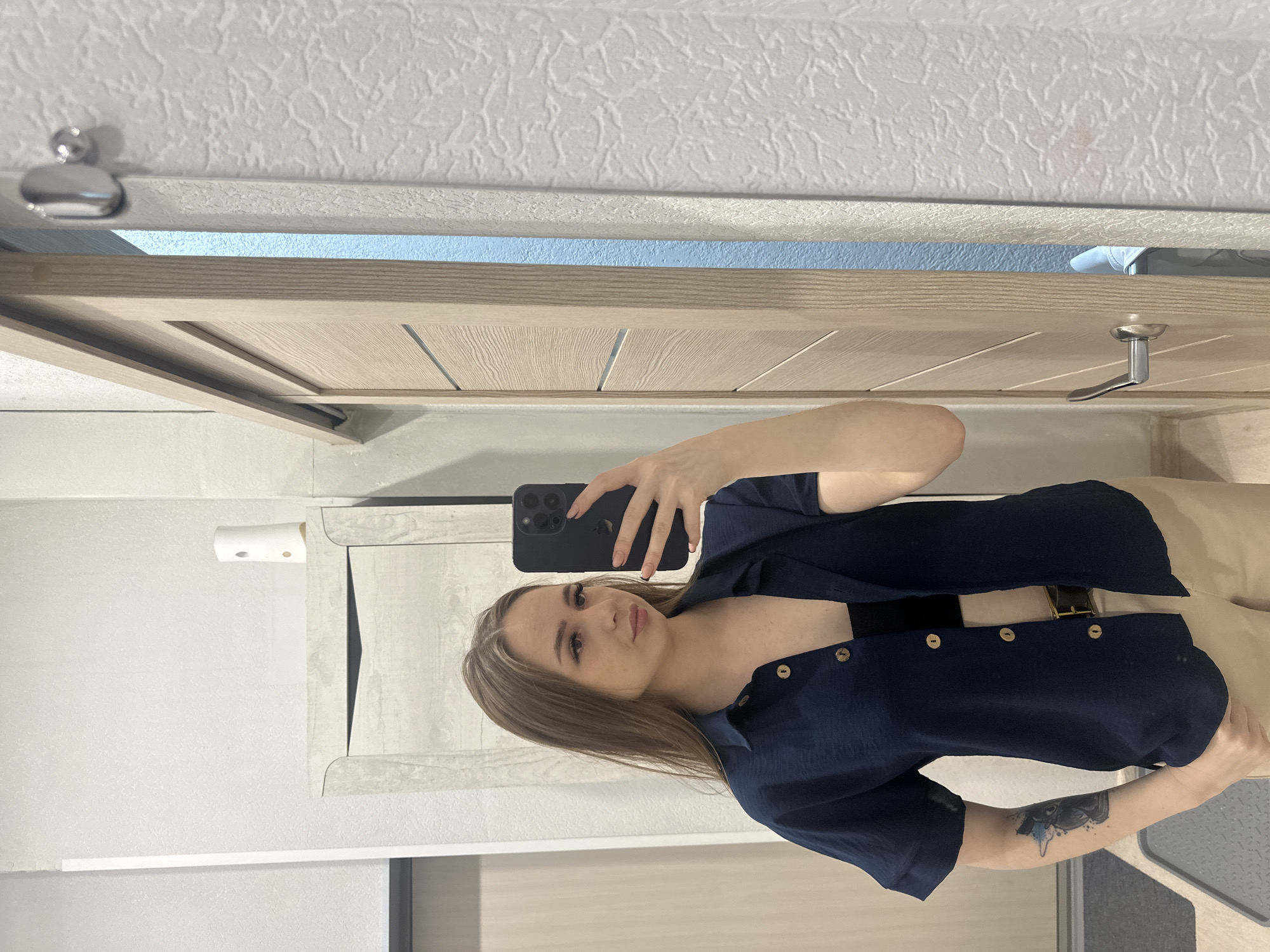 A woman with long, light brown hair is taking a selfie with a black smartphone. She is wearing a dark blue button-down shirt, with some of the buttons undone to reveal a black top underneath. She is also wearing light tan pants and a belt. A tattoo is visible on her left arm. The background consists of a bathroom setting, including a textured white wall, a light wood-toned door, and a toilet paper holder. The location appears to be a private residence, with no specific indication of a city or state.