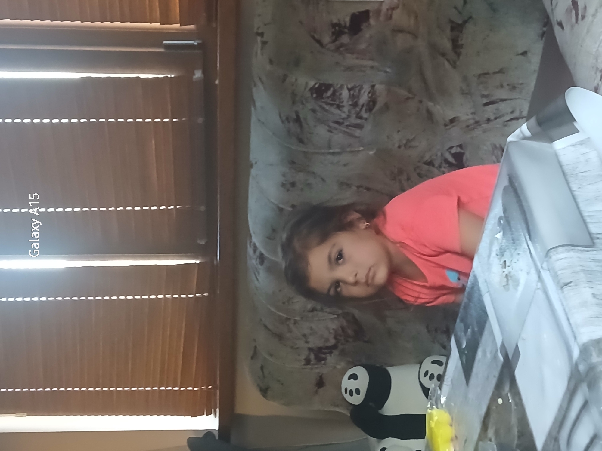A young girl in a pink shirt lies on a gray patterned couch next to a white table with a reflective surface. Beside her is a plush panda bear. A window with closed blinds is in the background. The location is assumed to be Obrochishte, Bulgaria.